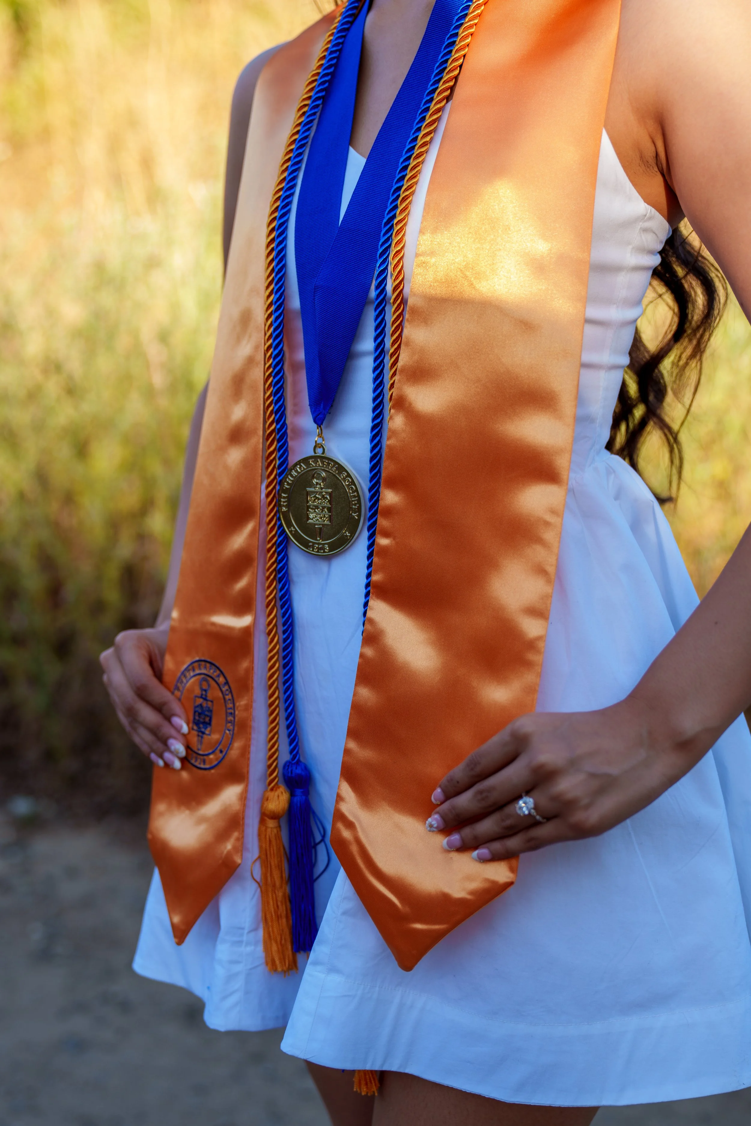 Close-up of a woman wearing a white dress and orange graduation stole with blue and gold cords, and a gold medallion around her neck, outdoors.