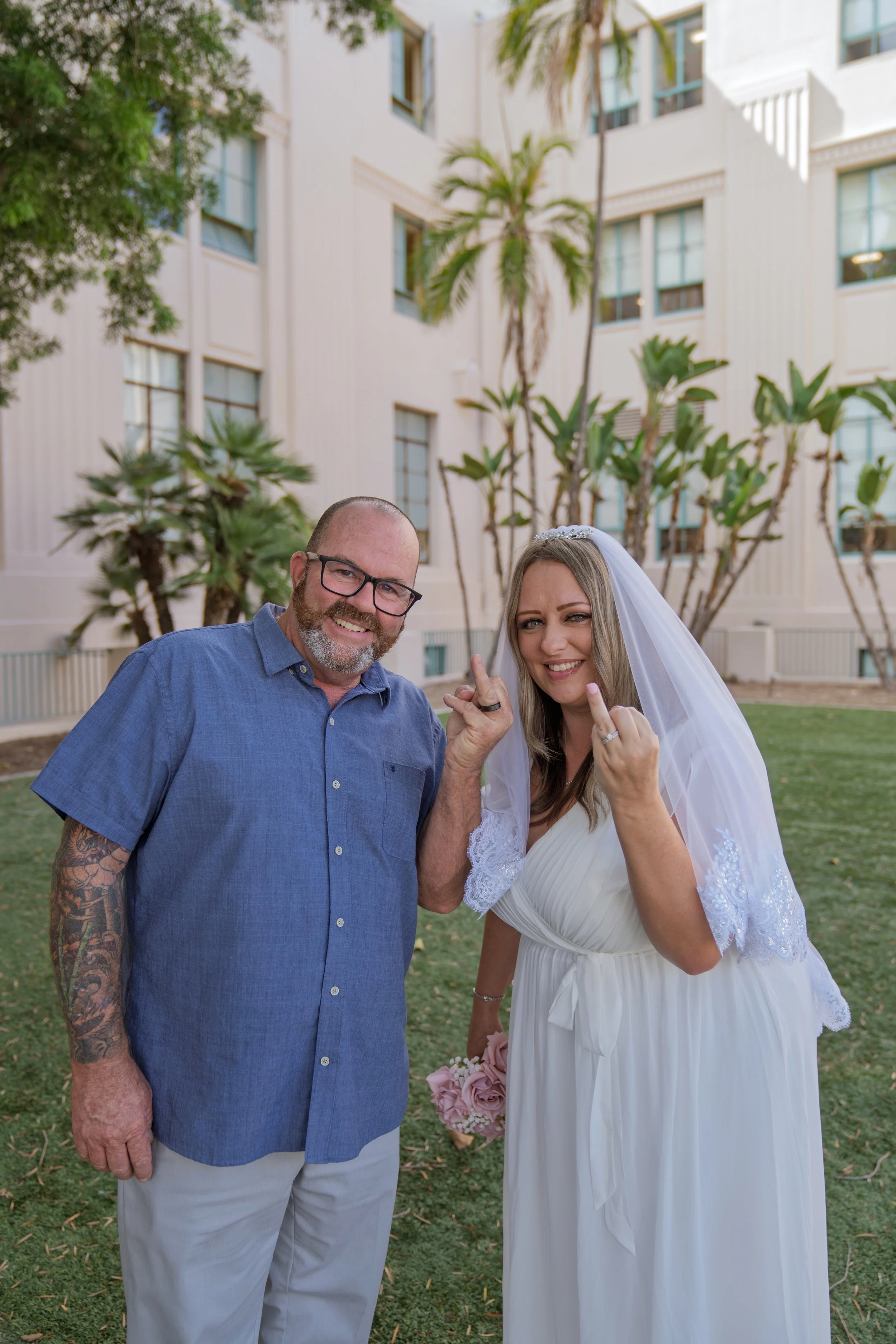 A man with glasses, a beard, and tattoos on his arm, smiling, standing next to a woman in a white wedding dress and veil, both making a gesture with their hands in an outdoor setting with green grass, trees, and a white building in the background.