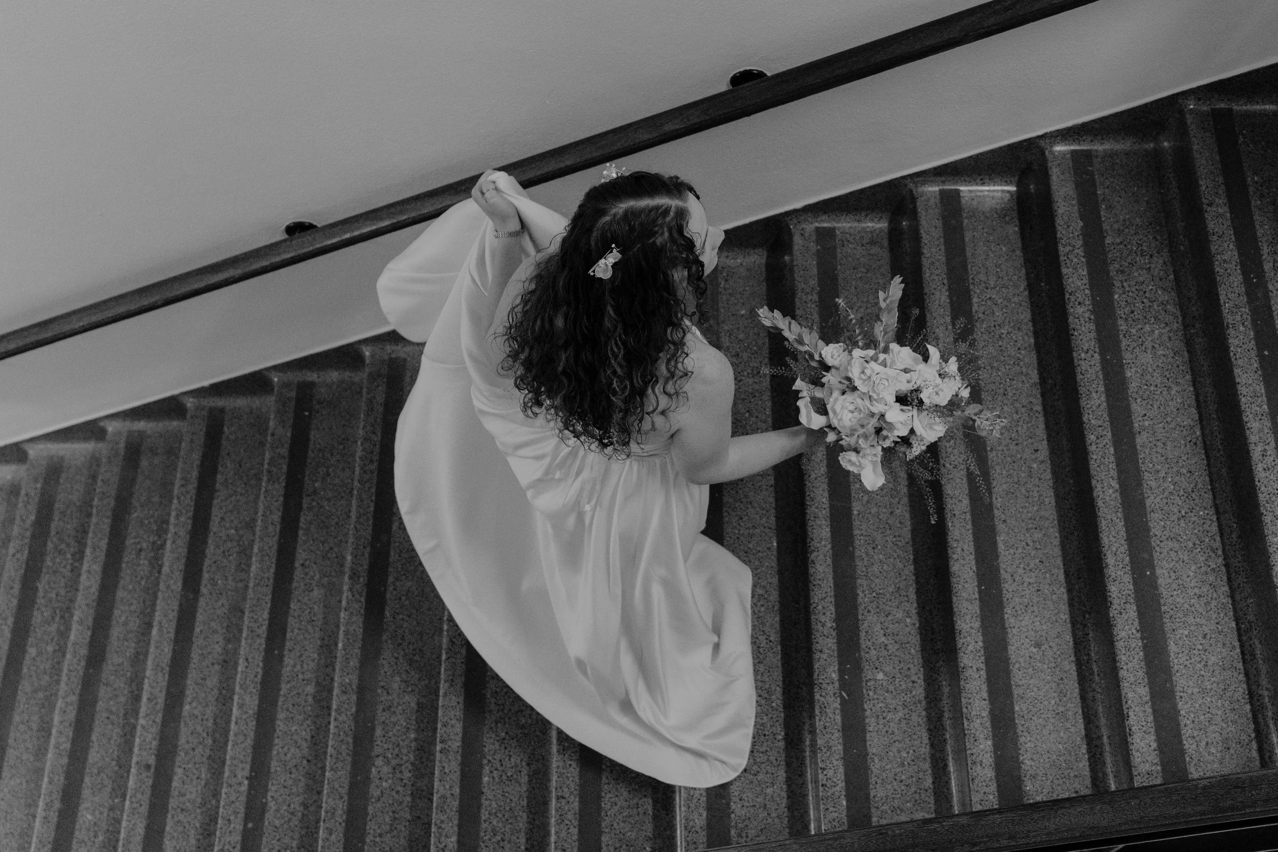 A woman in a wedding dress holding a bouquet of flowers walking down a staircase, viewed from above.
