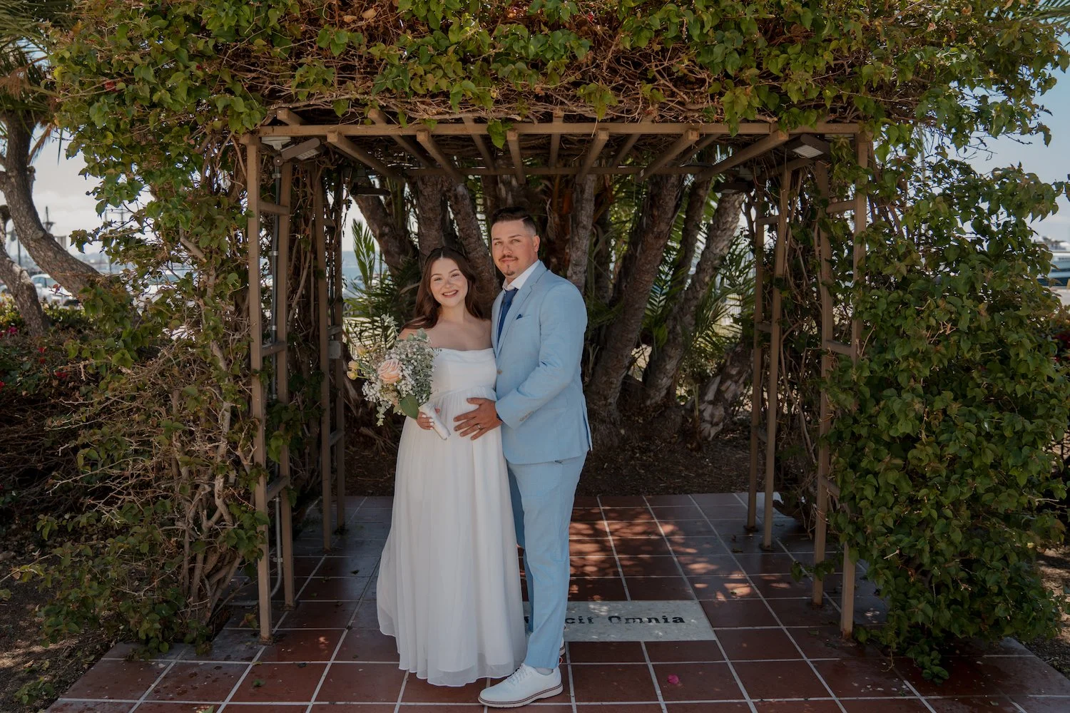 A bride and groom standing under a wooden arch covered with green foliage on a sunny day. The bride is holding a bouquet of flowers, wearing a white strapless wedding dress, and the groom is dressed in a light blue suit with white shoes. They are smi