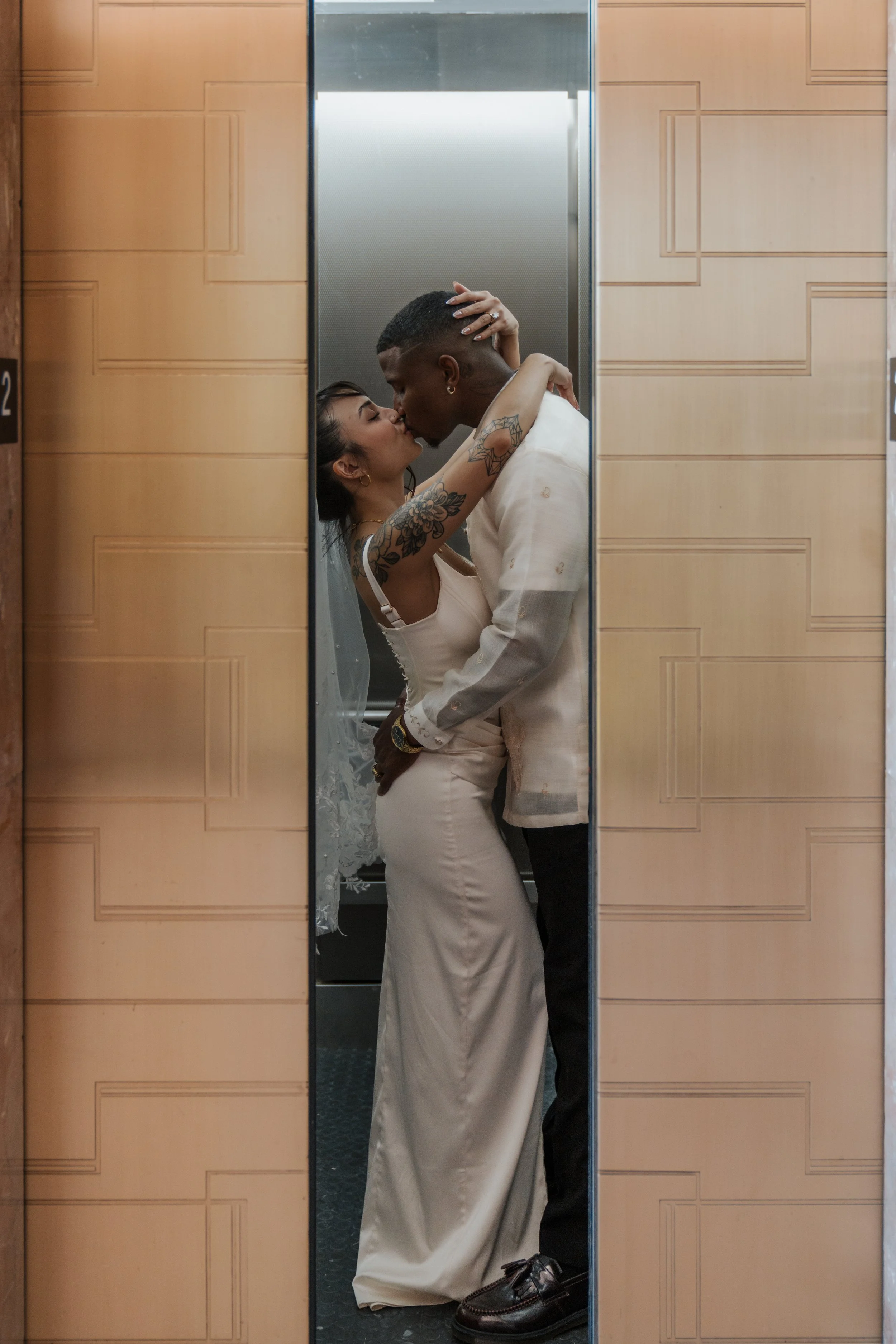 A couple is kissing in an elevator, with the woman wearing a white dress and the man wearing a light-colored suit. Their reflection is visible in the mirror of the elevator.
