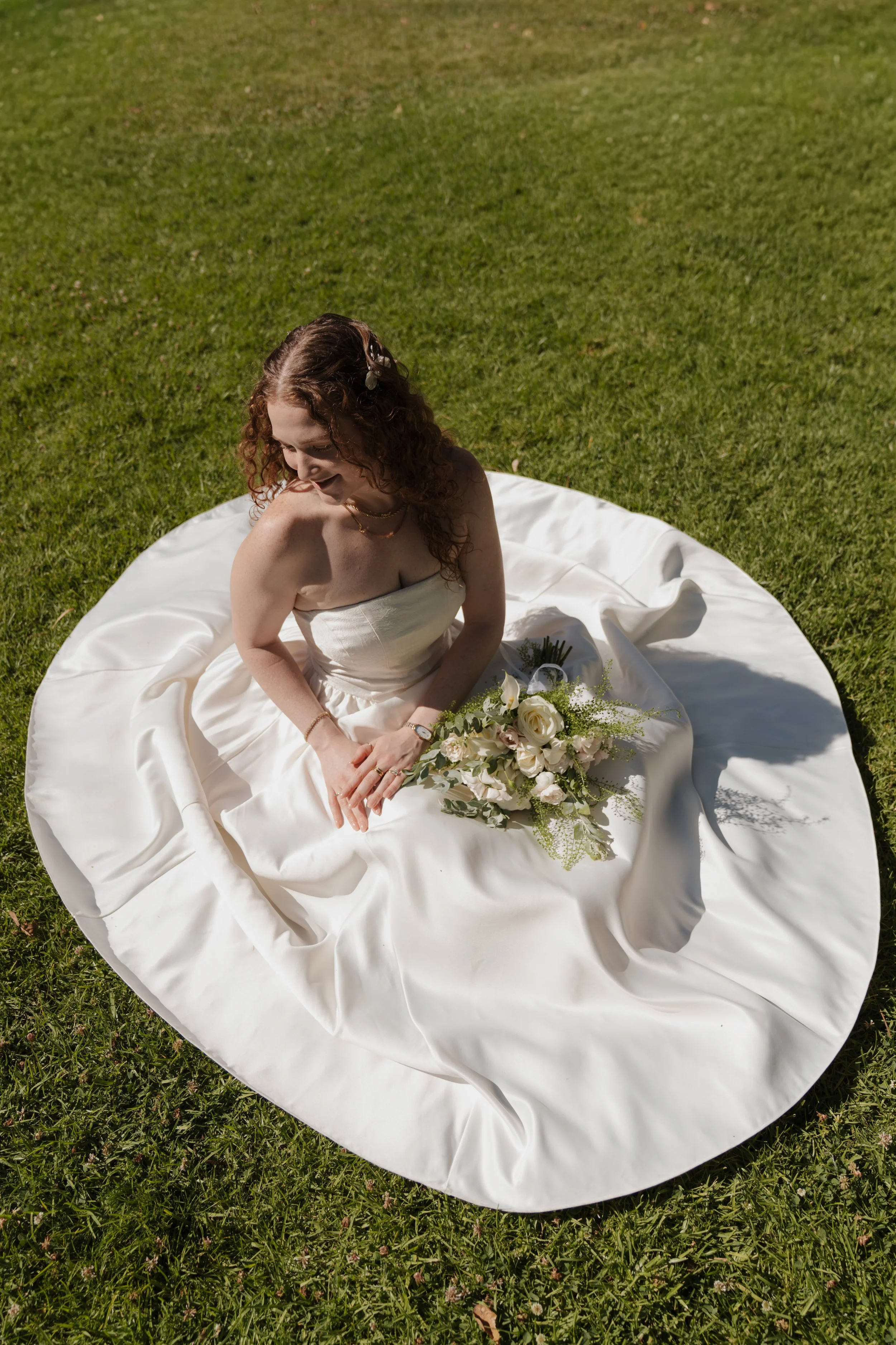 A woman in a white strapless dress sitting on a white round fabric on grass, holding a bouquet of white flowers with green foliage.