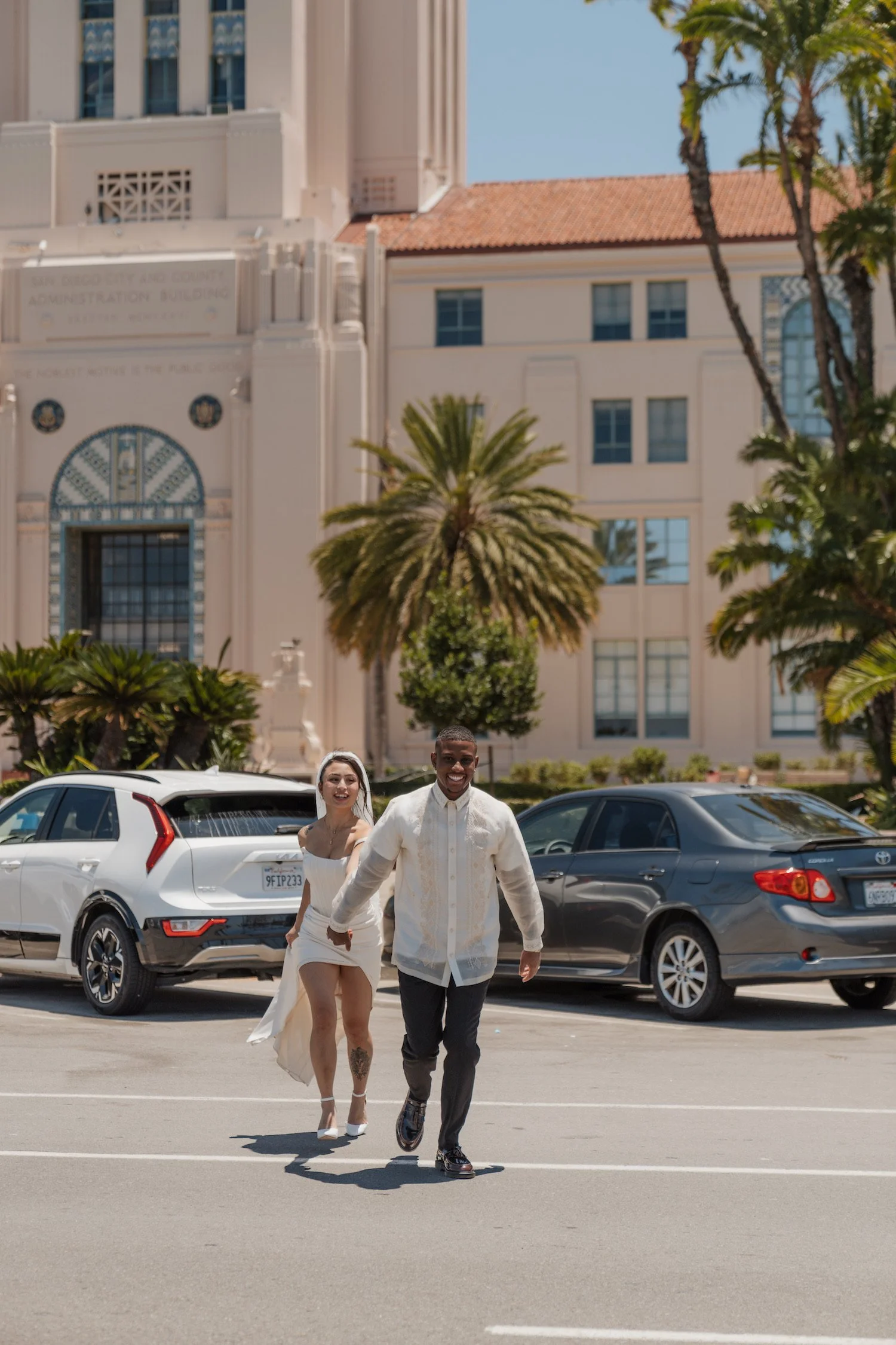 Young couple walking and smiling in a parking lot with cars and palm trees in the background, in front of a large building.