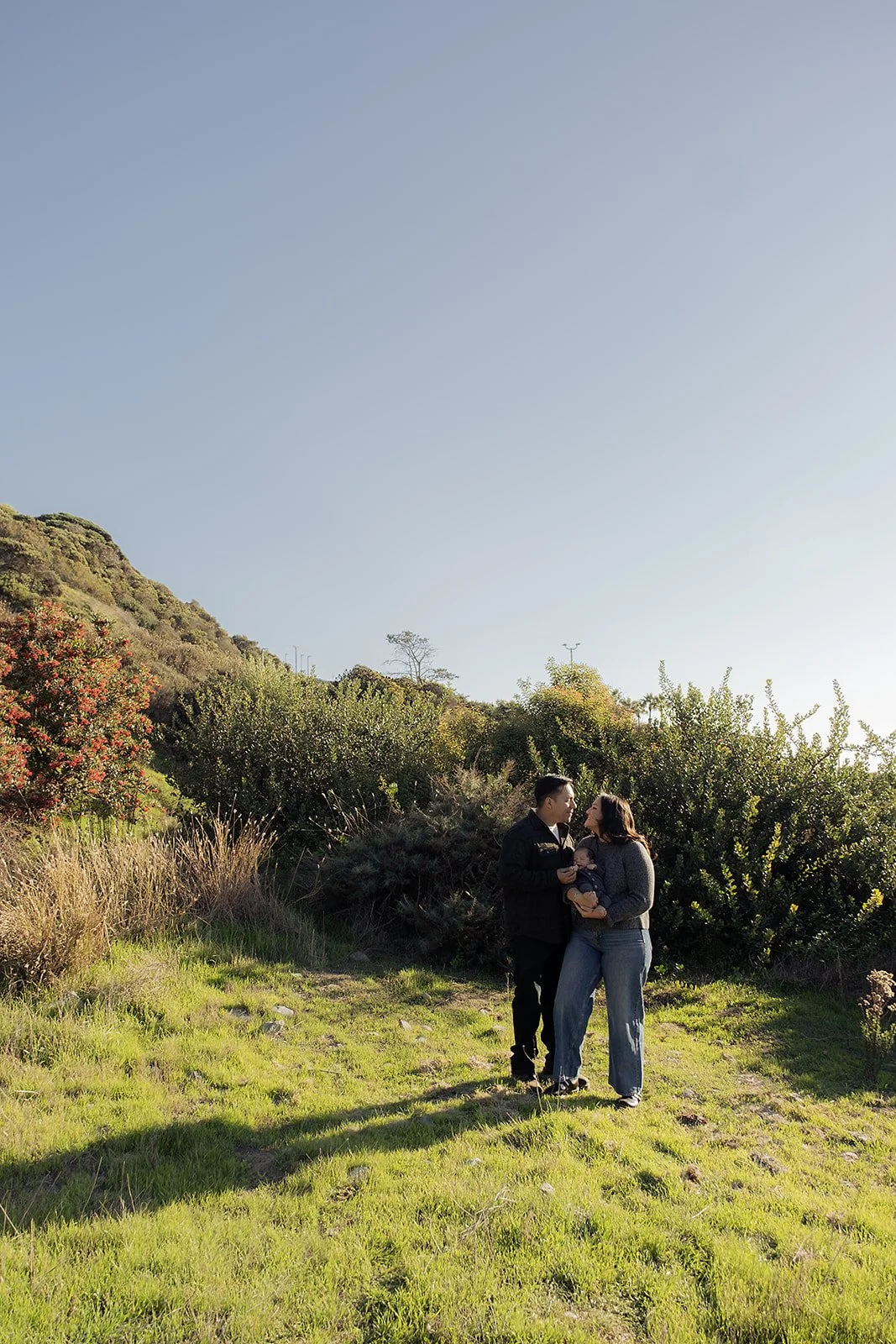 A family of three standing on a grassy hillside with trees and bushes, under a clear blue sky.