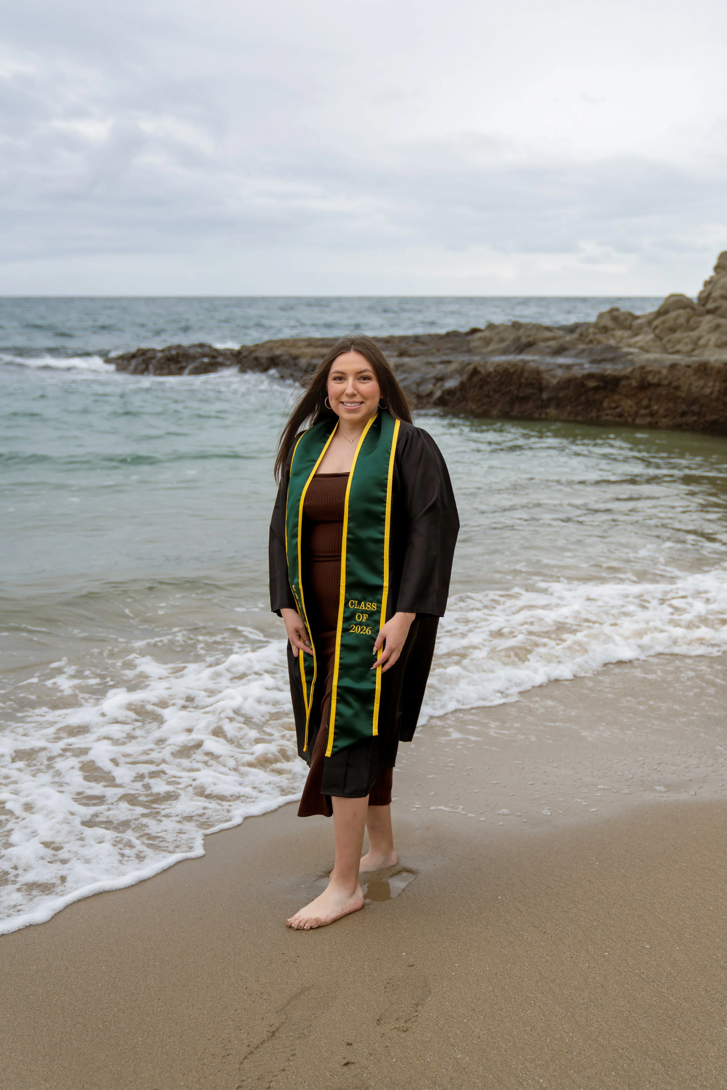 A young woman in a black graduation gown with a green and gold stole, standing on a sandy beach with waves and rocks in the background, smiling at the camera.
