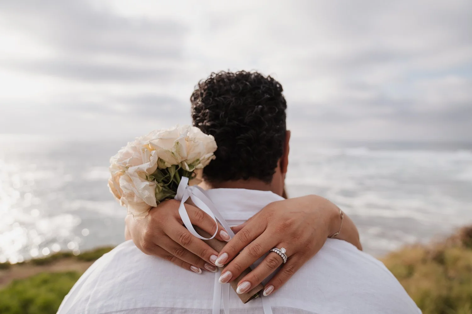 A couple hugging by the ocean, with the woman holding a bouquet of white roses and showcasing an engagement ring.