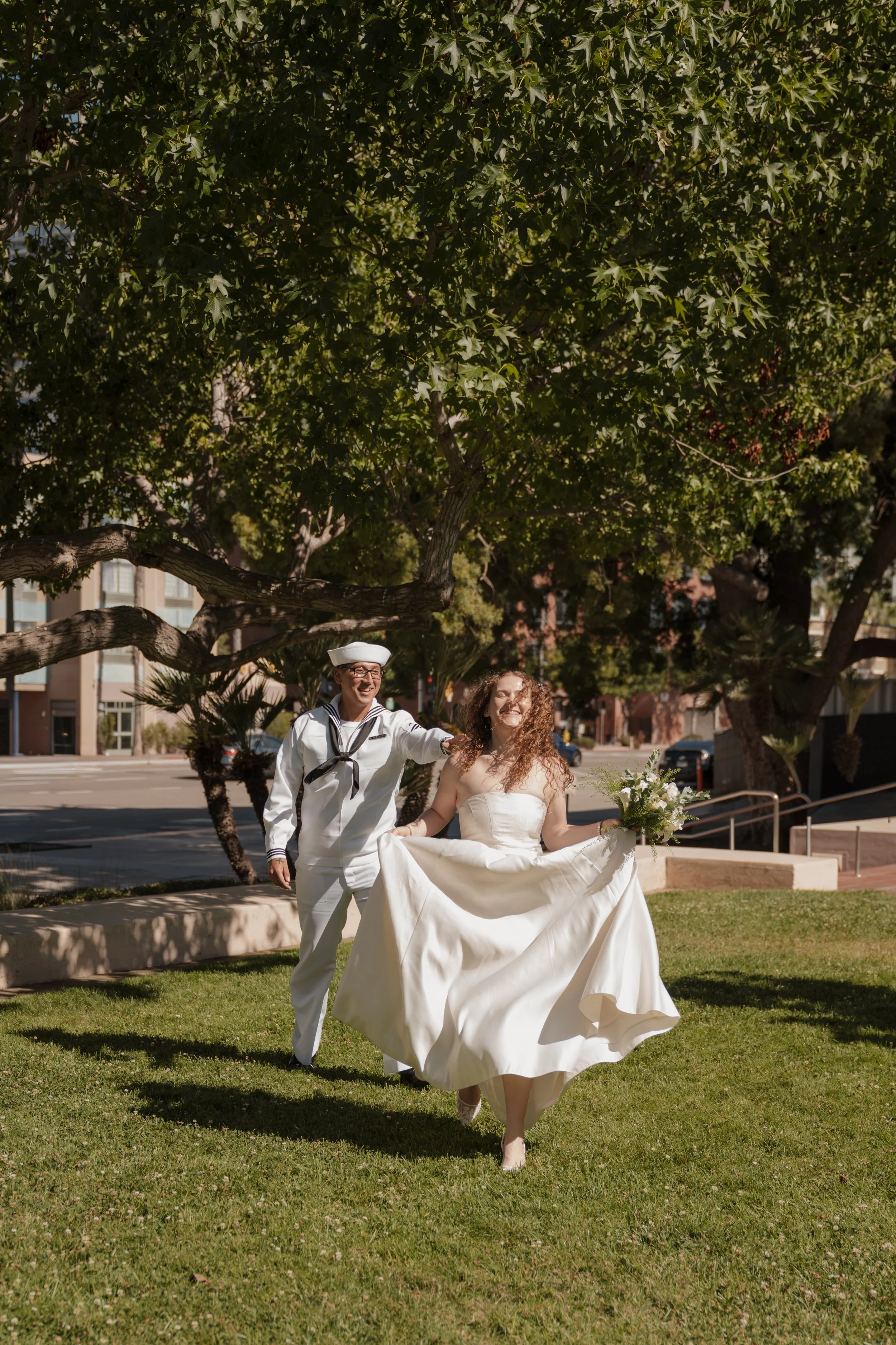 A couple dressed in wedding attire walking on a grassy area under a large tree in an urban park, with buildings in the background. The bride is holding a bouquet, and the groom is wearing a sailor suit.