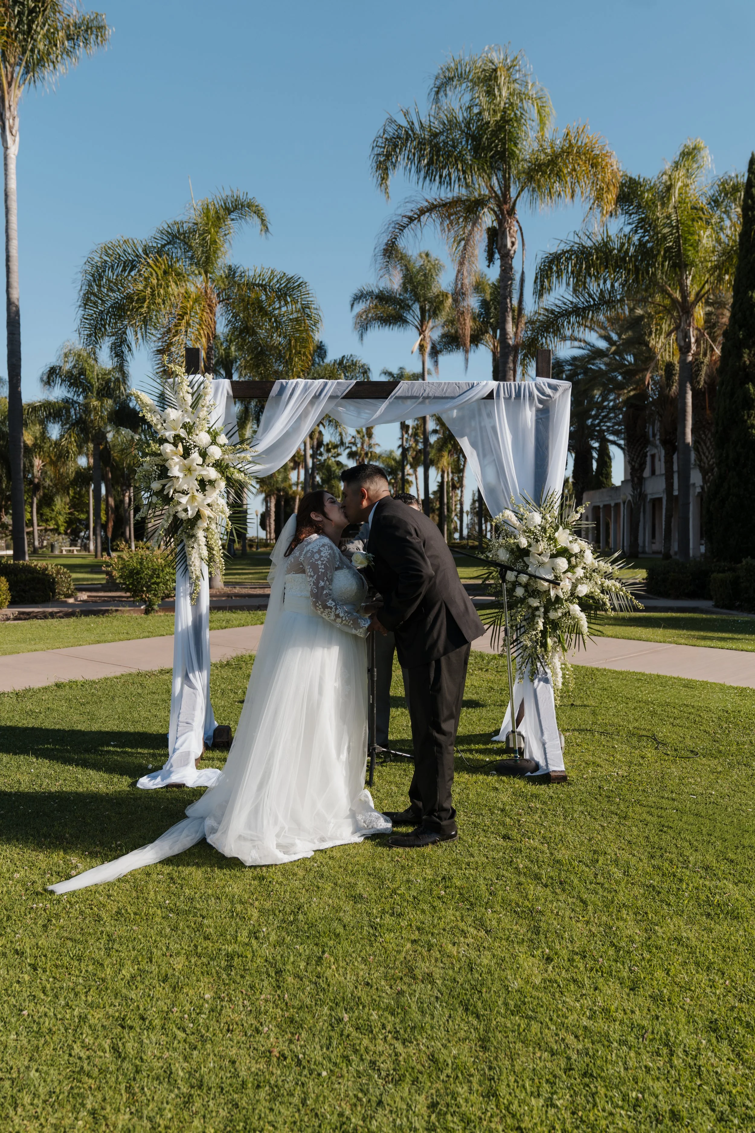A bride and groom kissing during their outdoor wedding ceremony under a decorated arch with white drapery and floral arrangements, with palm trees and clear blue sky in the background.