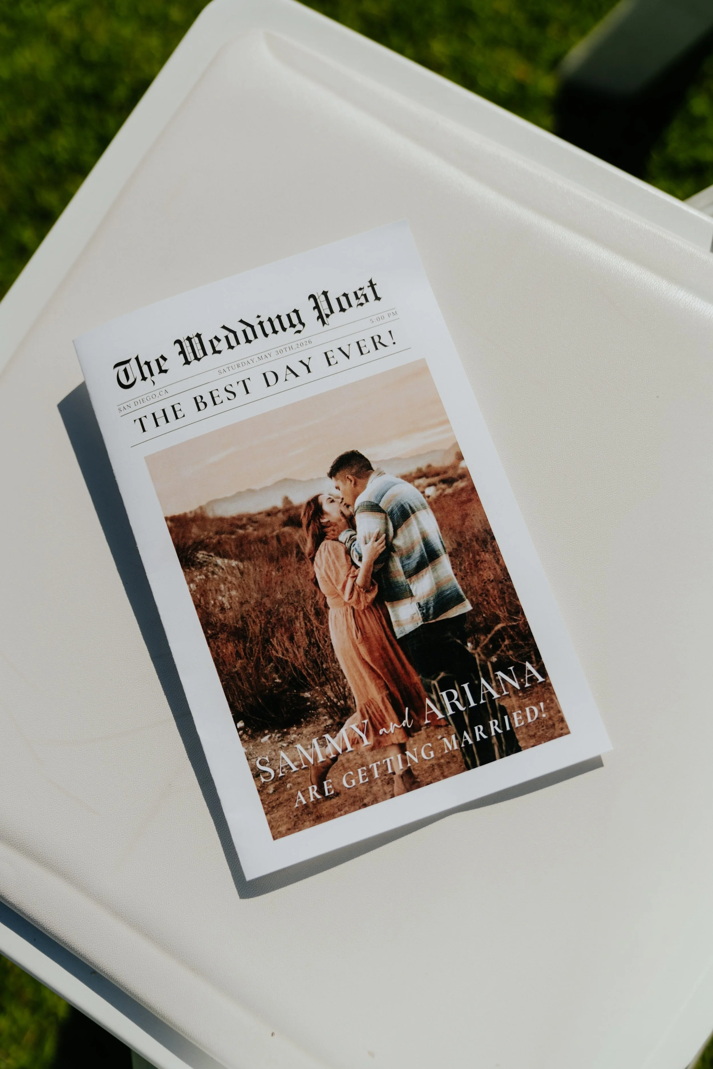 A wedding program flyer titled 'The Wedding Post' on top of a white surface outdoors with grass in the background. The flyer features a photo of a couple, a man and a woman, standing in a field, embracing and smiling at each other.
