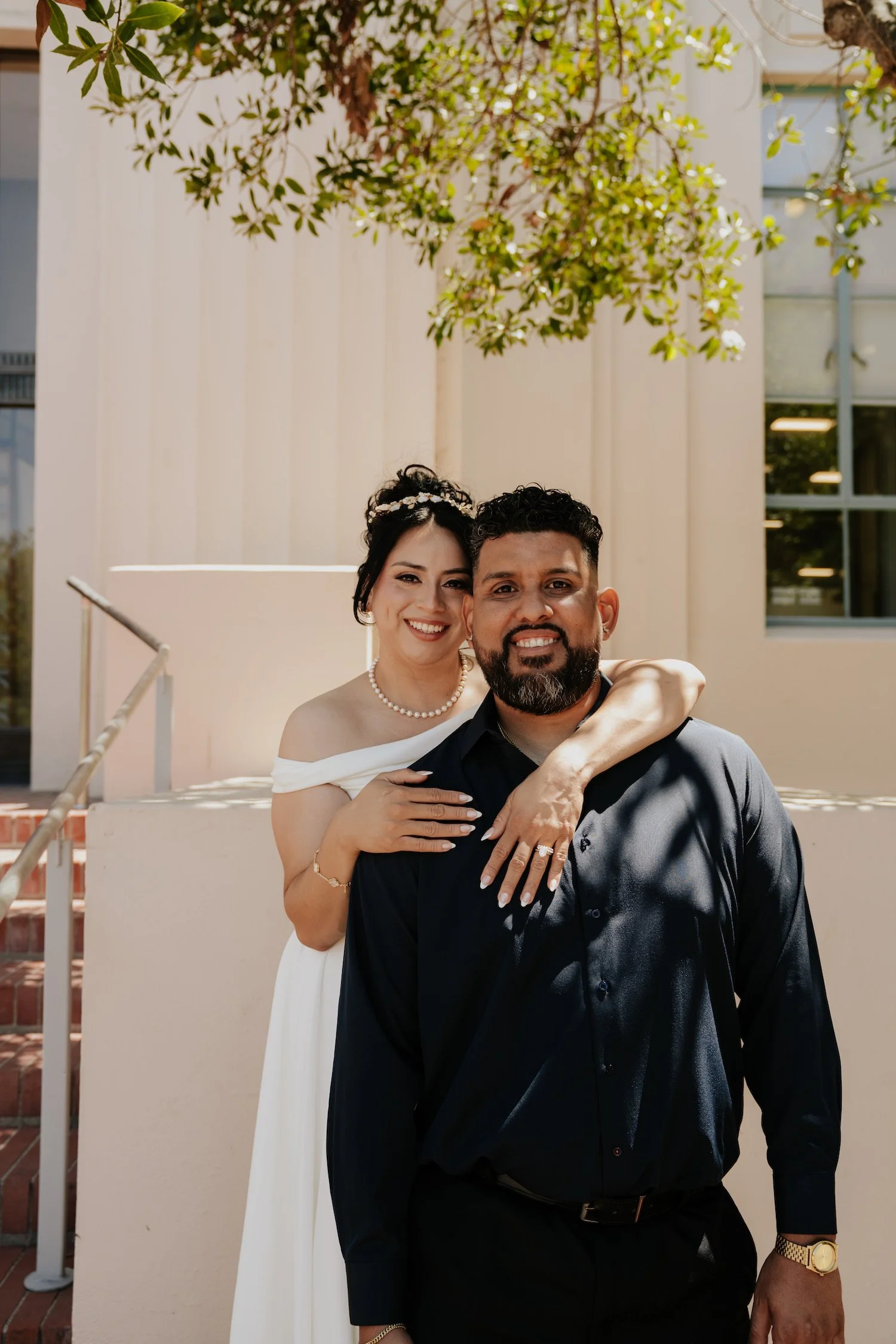 A smiling woman with dark hair styled in an updo, wearing a white off-shoulder dress, pearl necklace, and bracelet, stands behind a man with dark curly hair and a beard, dressed in a black shirt and pants, outdoors in front of a beige building with w