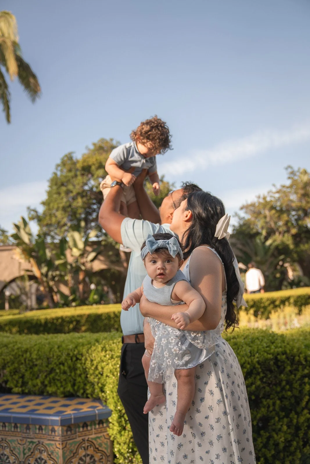 A family of four outdoors on a sunny day, with a woman holding a baby girl with a headband, a man lifting a young boy, and trees and a hedge in the background.