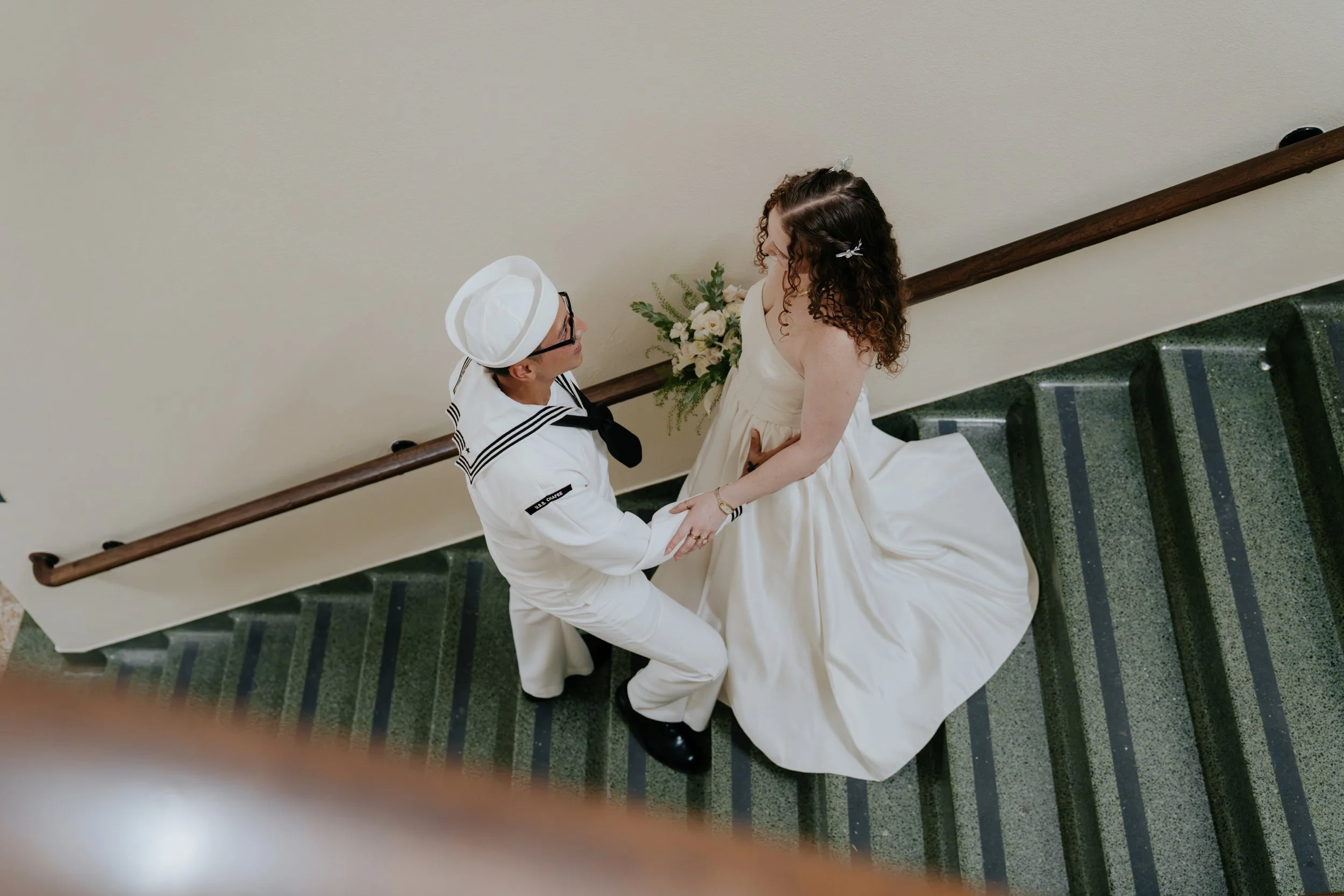 A person dressed as a sailor kneeling before a woman in a cream-colored dress on a staircase, with a bouquet of flowers in the background.