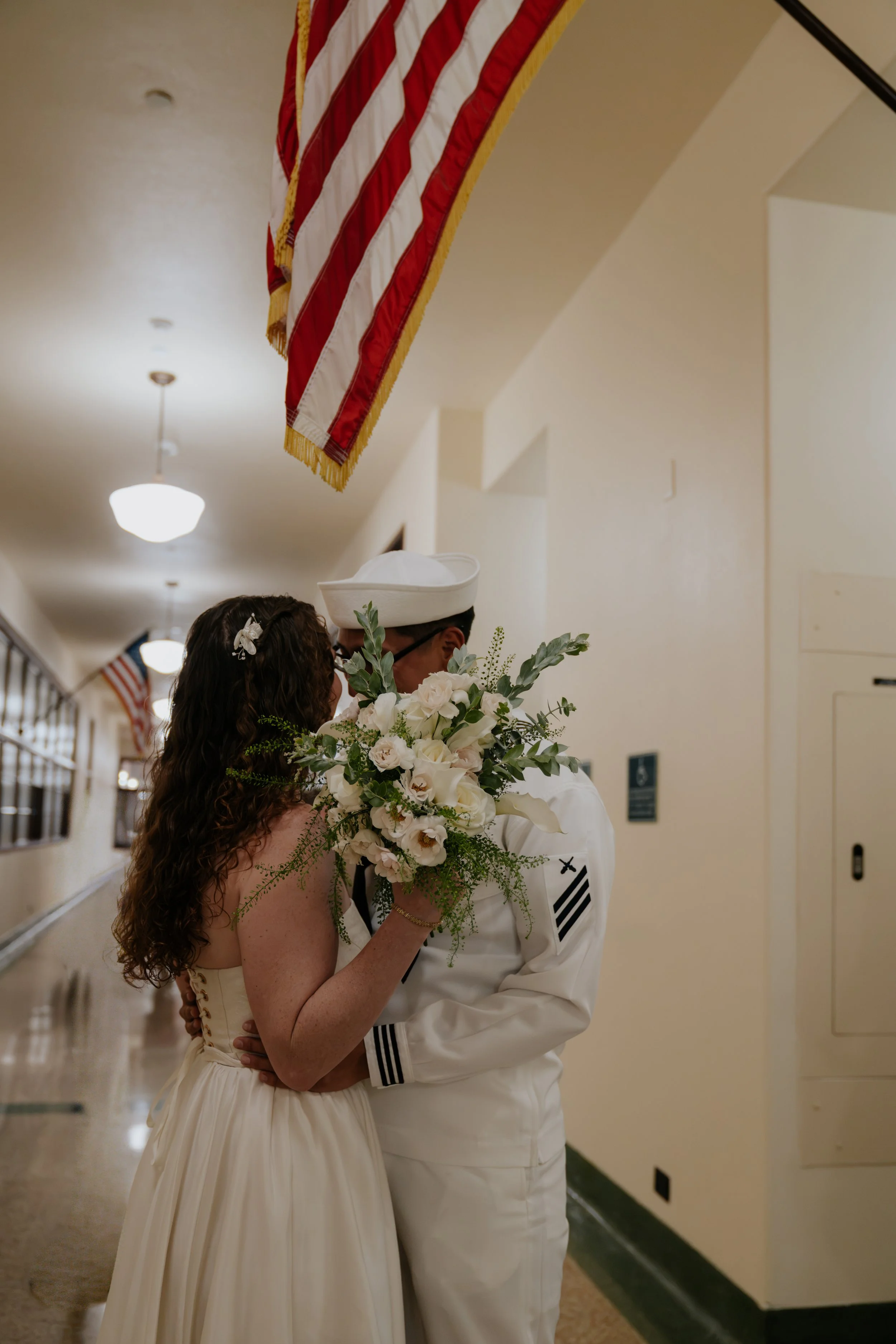 A bride and groom sharing a kiss indoors, with American flags hanging from the ceiling in the background. The bride has long, curly hair and holds a bouquet of white flowers. The groom is in a military uniform, and they are embracing closely.