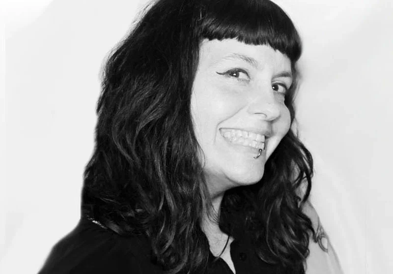 Black-and-white photo of a woman with dark, wavy hair, smiling and showing a side profile with a lip piercing.