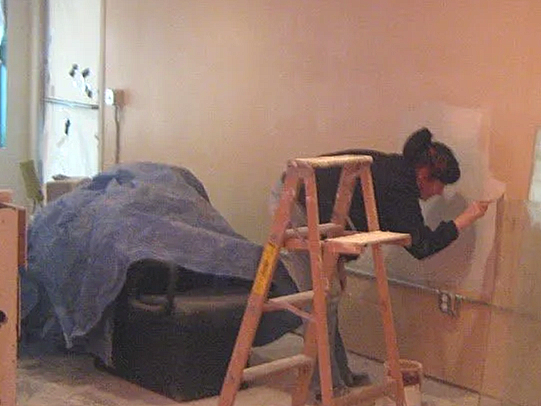A person wearing a black jacket and cap working on the wall near a ladder in a room with a bed.