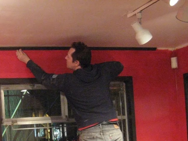 A person standing on a surface, reaching up to install or adjust something along the ceiling in a room with red walls and track lighting.