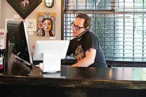 Man with glasses working at a desk with a computer and a phone, in a cafe setting.