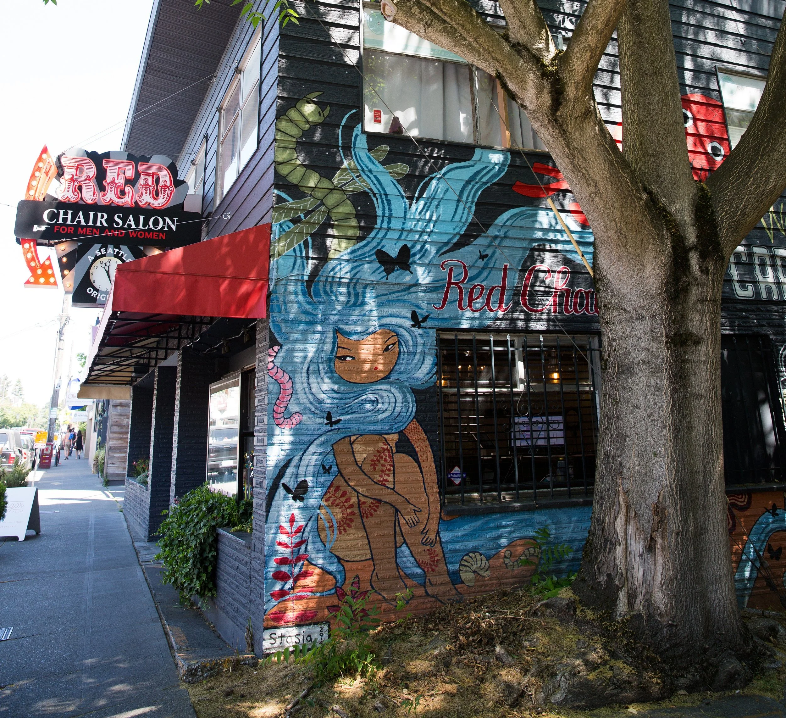 Colorful mural of a girl with long blue hair, surrounded by black butterflies, on the side of a building. There is a tree in front and a sign for a chair salon nearby.