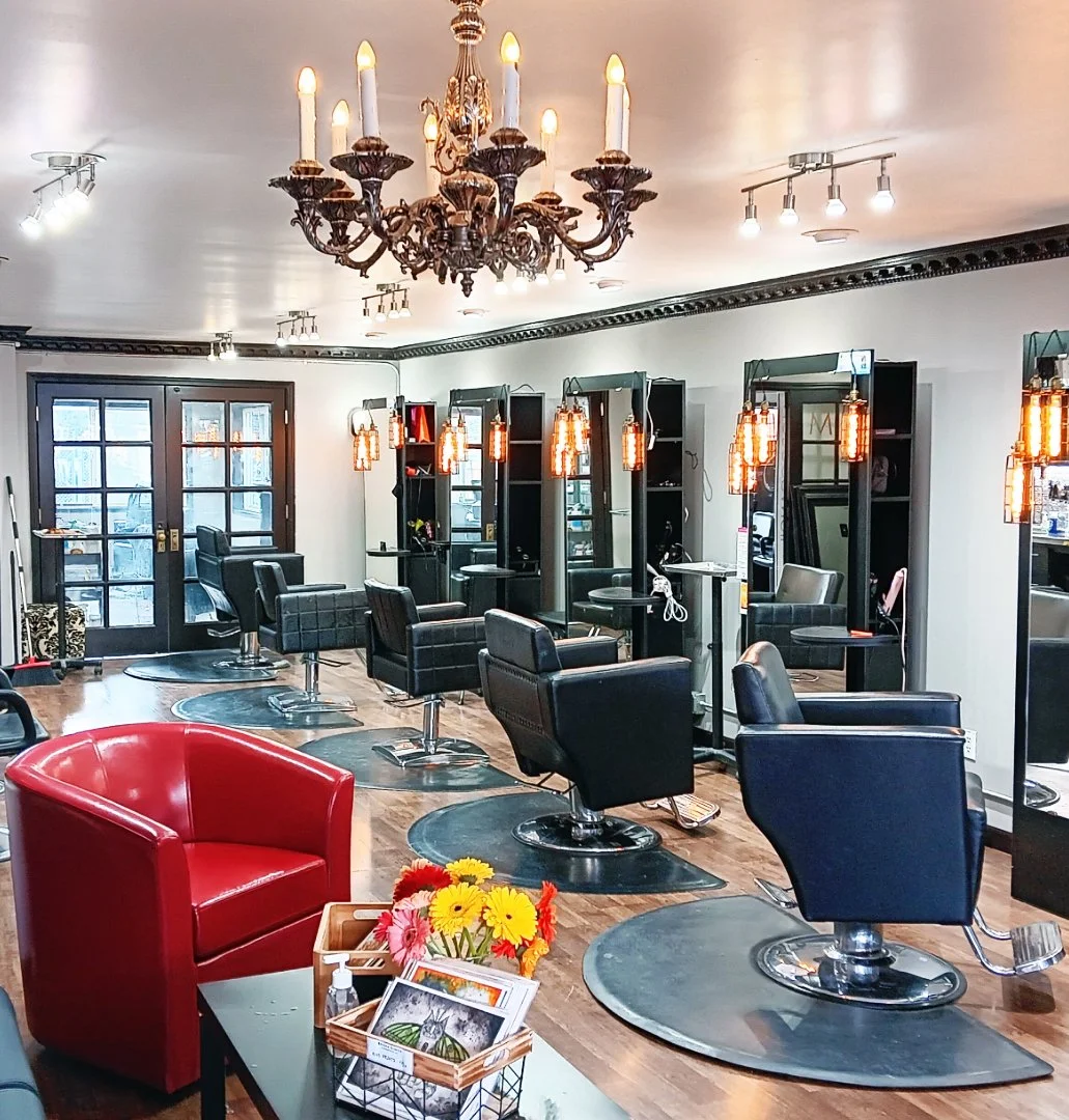 An interior view of a modern hair salon with black styling chairs, mirrors, and warm lighting fixtures. There is a red armchair and a small table with flowers and magazines in the foreground, and a chandelier hanging from the ceiling.