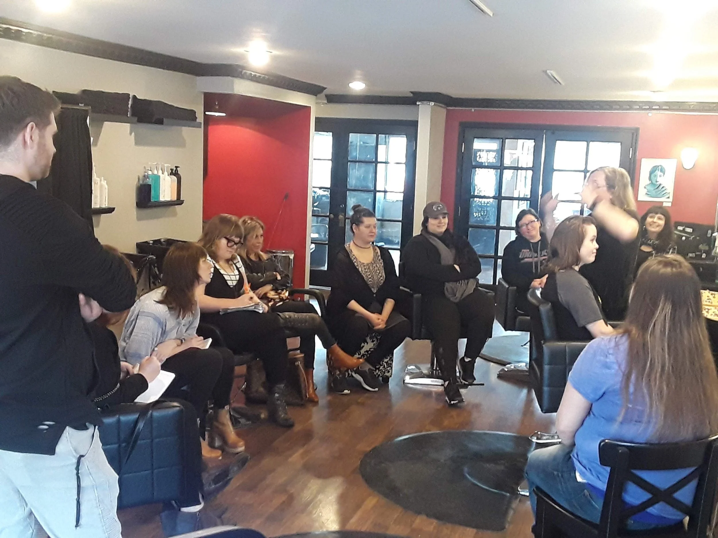 Group of women seated in a salon or community room, with a woman standing and speaking, surrounded by hair products on shelves, and large windows in the background.