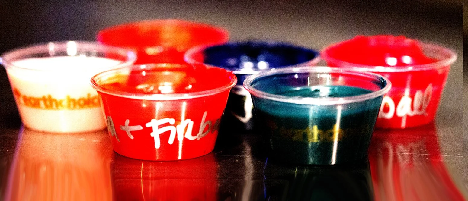 Colorful tealight candles in small plastic cups with the logo 'Fireball' printed on them, placed on a reflective surface.