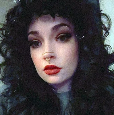 A woman with black hair, heavy eye makeup, dark lipstick, and facial piercings, wearing a black spaghetti strap top, in front of a plain wall.