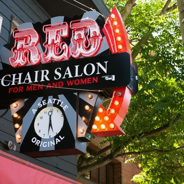 A neon sign for a chair salon with the words 'REO Chair Salon for Men and Women' and a clock indicating 12:05, mounted on a building with trees in the background.
