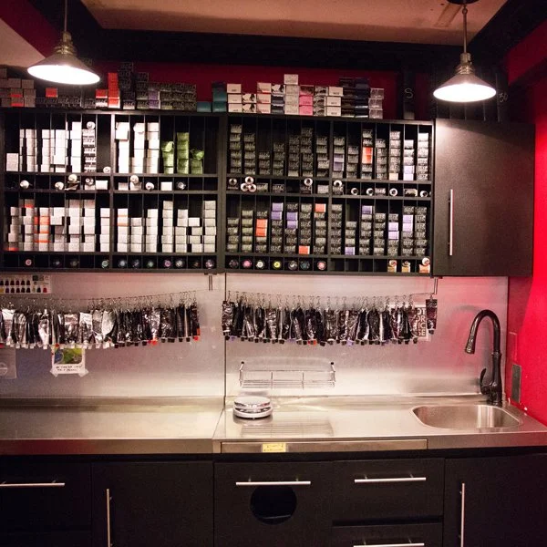 A retail workspace with black wall-mounted shelves filled with colorful boxes and rolls, hanging black plastic bags, and a stainless steel countertop with a sink and faucet.