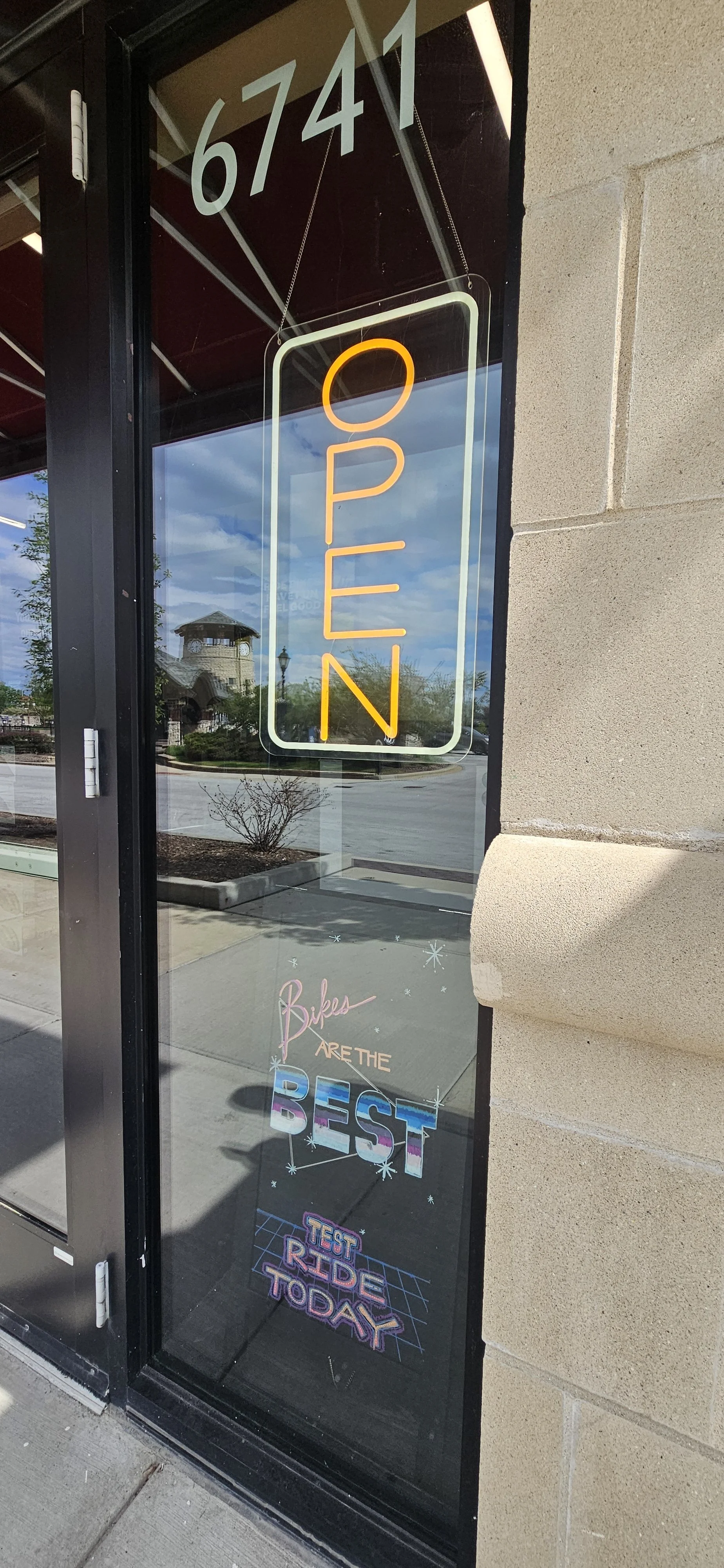Storefront window with a neon "OPEN" sign hanging. Inside the window, a colorful sign says "Bikes ARE THE BEST" and a neon sign reads "TEST RIDE TODAY." Reflection of a building and sky outside.