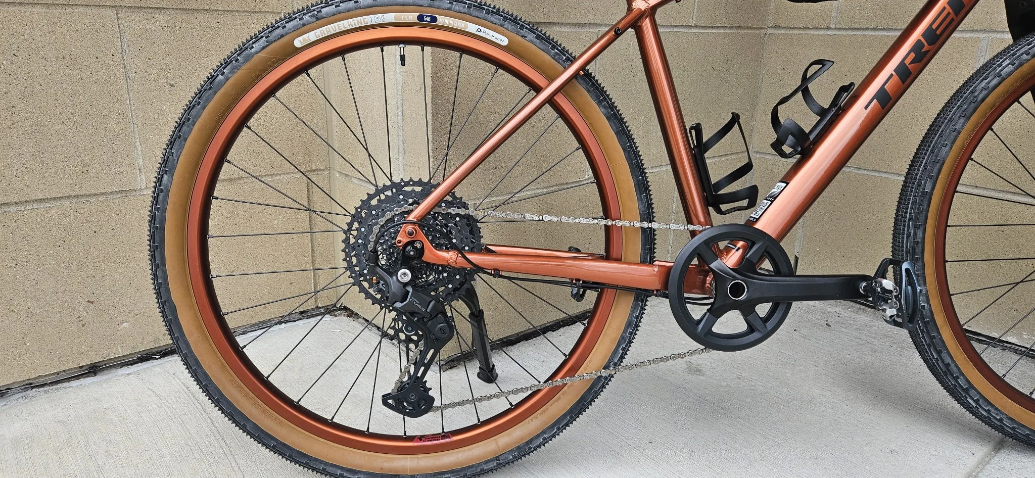 A close-up of a mountain bike with a metallic brown frame, tan tires, and disc brakes, leaning against a concrete wall.