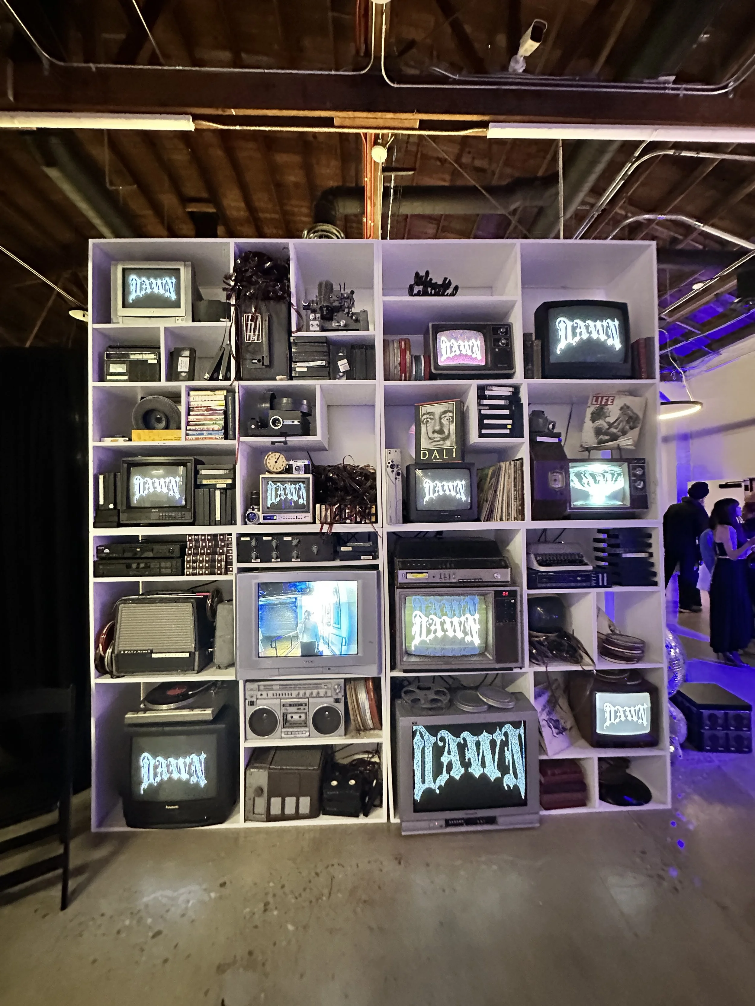 A display of vintage electronics and televisions, arranged on a white cubical shelving unit in an indoor setting with a brick ceiling and exposed pipes.