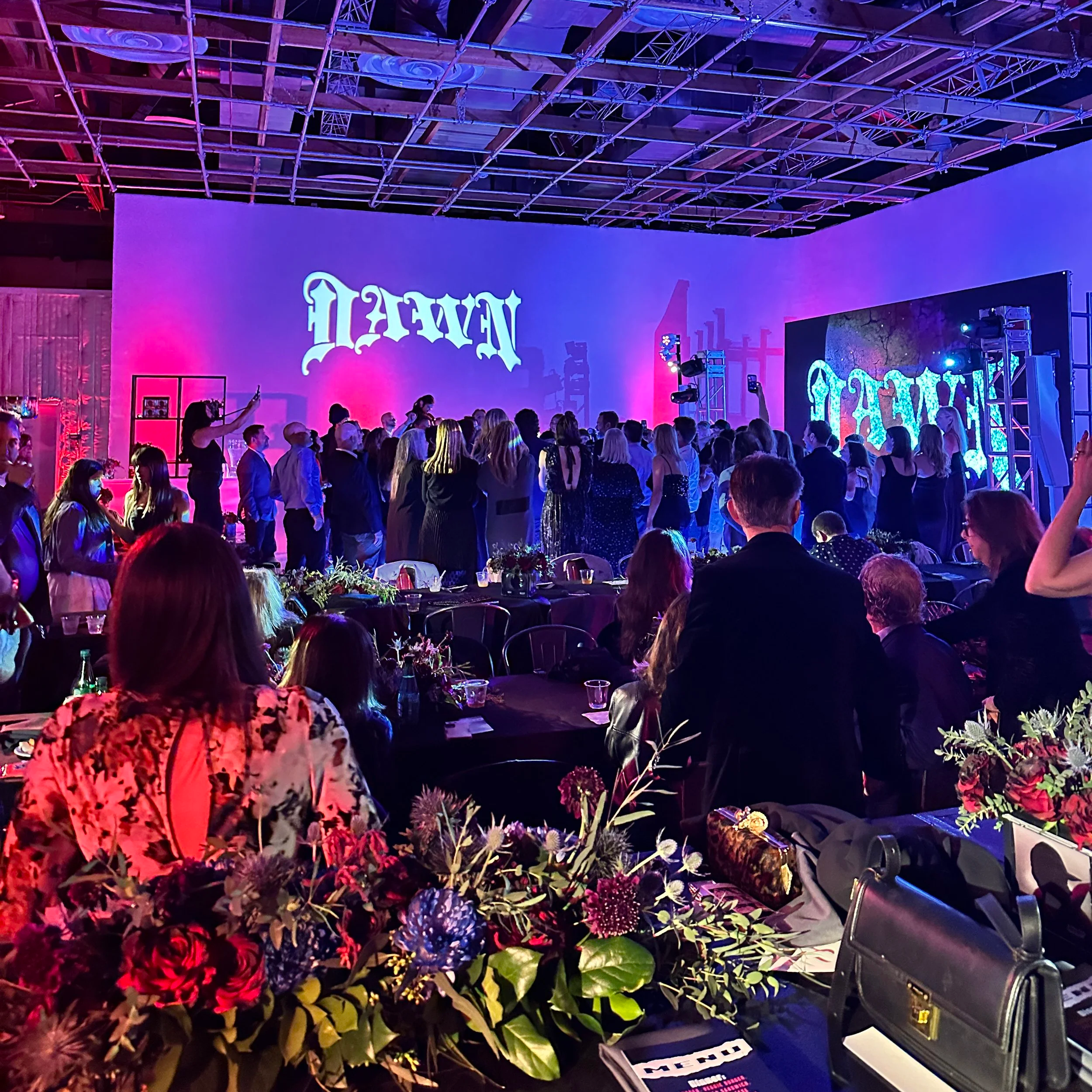 Indoor event with people gathered around tables, large floral arrangements, a projection displaying the word 'VAN' on a wall, and theater or stage lighting. The ceiling has a wooden framework with purple and pink lighting.