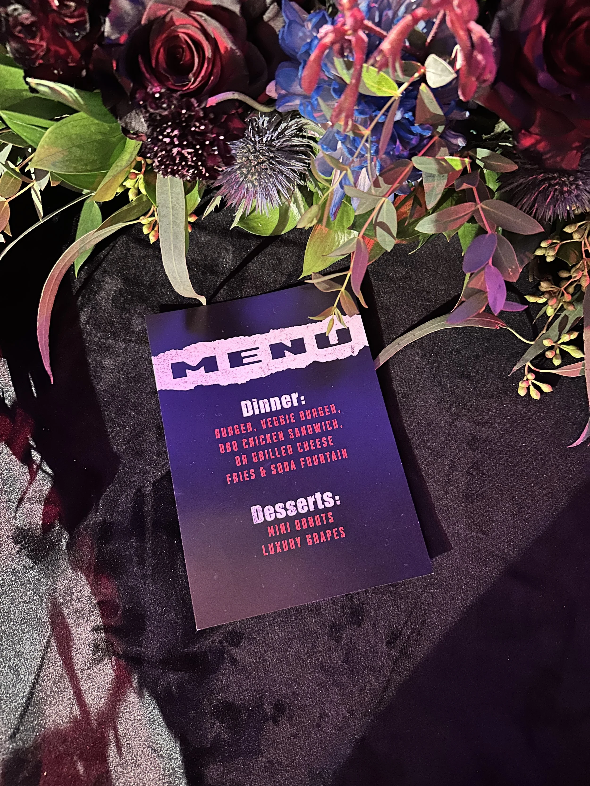 A photograph of a floral arrangement with red roses, purple and blue flowers, and green foliage, placed on black fabric. A menu card is partially visible, listing dinner options such as burger, veggie burger, BBQ chicken sandwich, or grilled cheese w