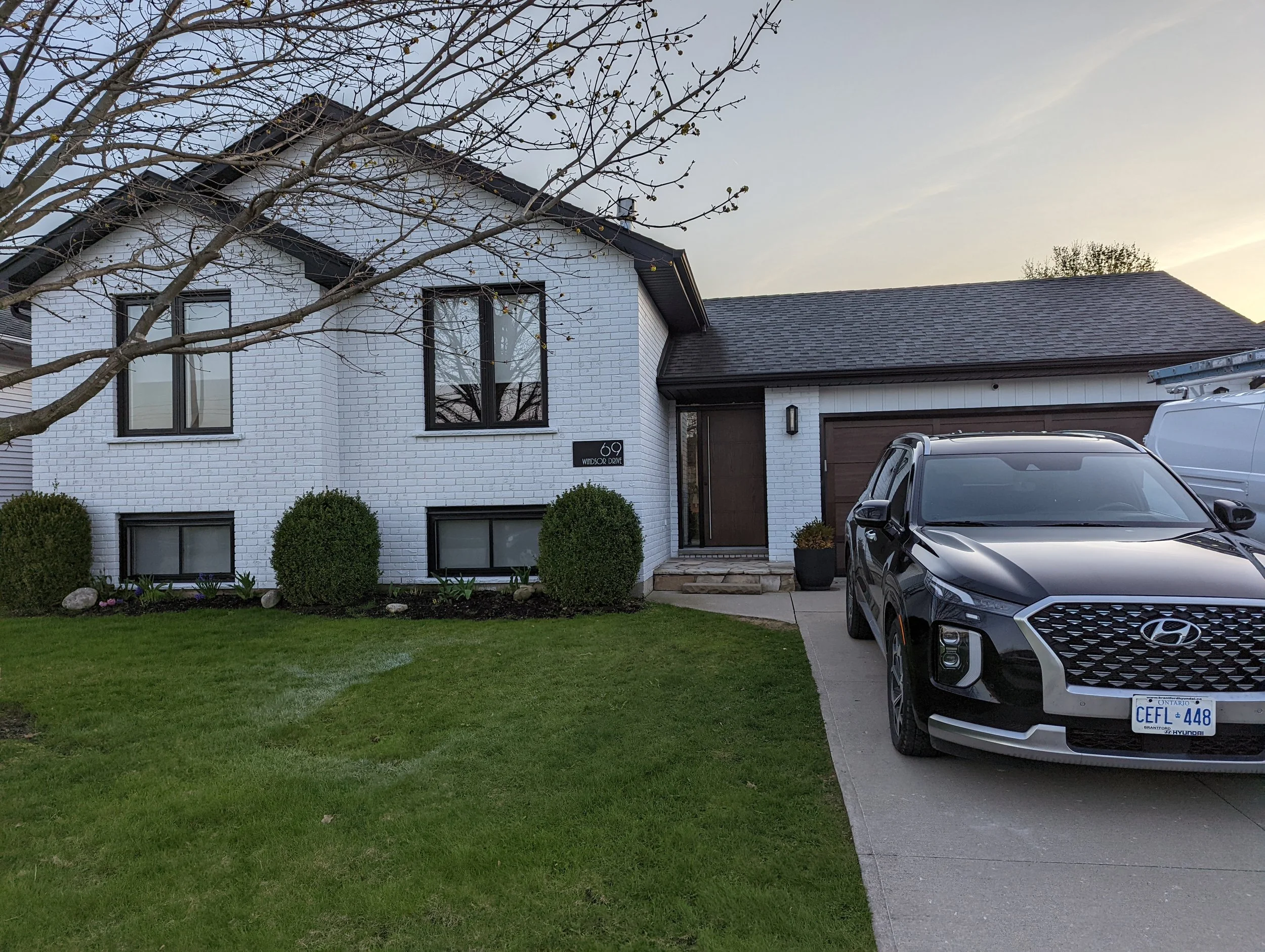 A white house with black window frames and a dark roof, a small front yard with green grass and bushes, a tree with bare branches in front, two parked cars, and a house number sign that reads "69 Windsor Drive."