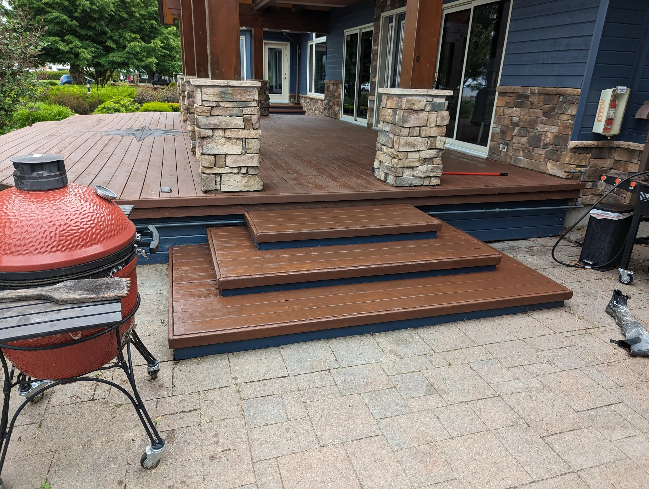 A backyard wooden deck with steps, supported by stone pillars, attached to a house with blue siding and large glass doors. There is a red ceramic grill in the foreground, a fire extinguisher mounted on the house wall, and landscaping with trees and bushes in the background.
