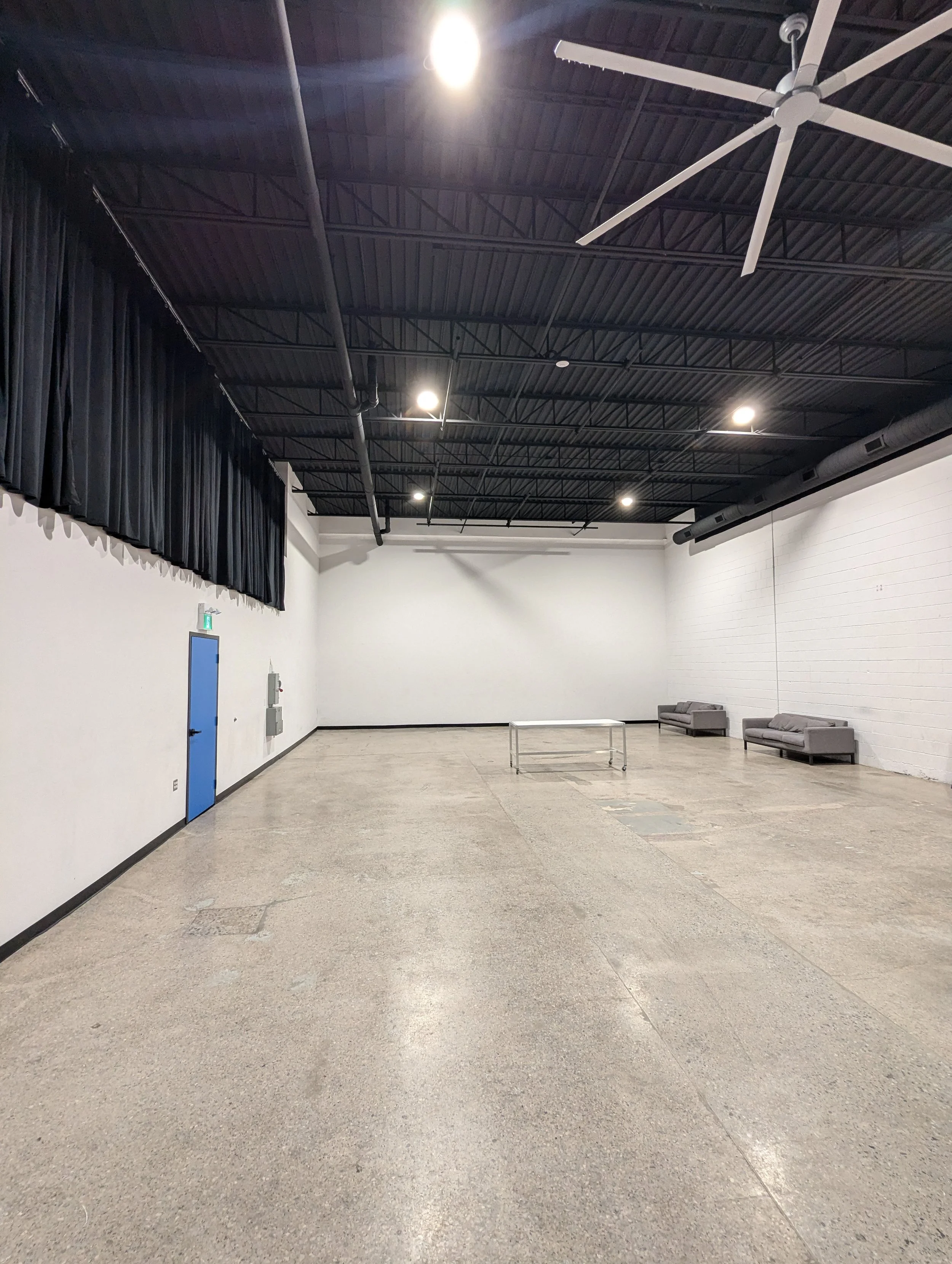 An interior space with a high, dark ceiling with exposed beams and industrial lighting. The walls are white, and there are a few gray couches and a small table along the walls. There is a blue door on the left side and a few electrical panels on the wall.