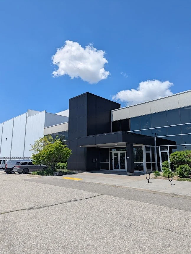 AModern commercial building with glass windows and a black entrance, parking lot with cars, green bushes, and a partly cloudy blue sky.