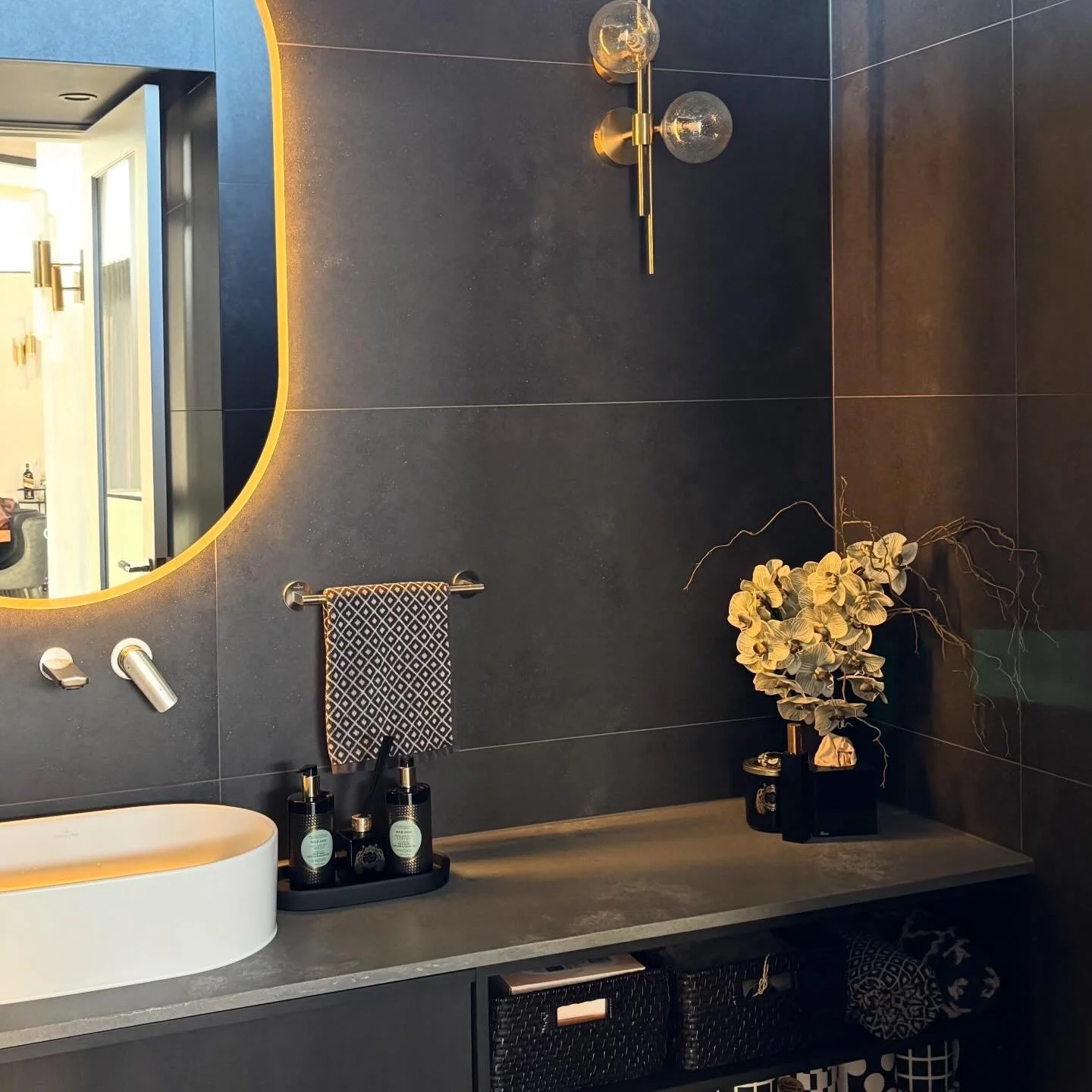 Modern bathroom vanity with a large round mirror, a black countertop with a white vessel sink, black bottles of soap or lotion, a decorative plant in a black vase, a beige towel with a black geometric pattern, and wall-mounted light fixtures with sph