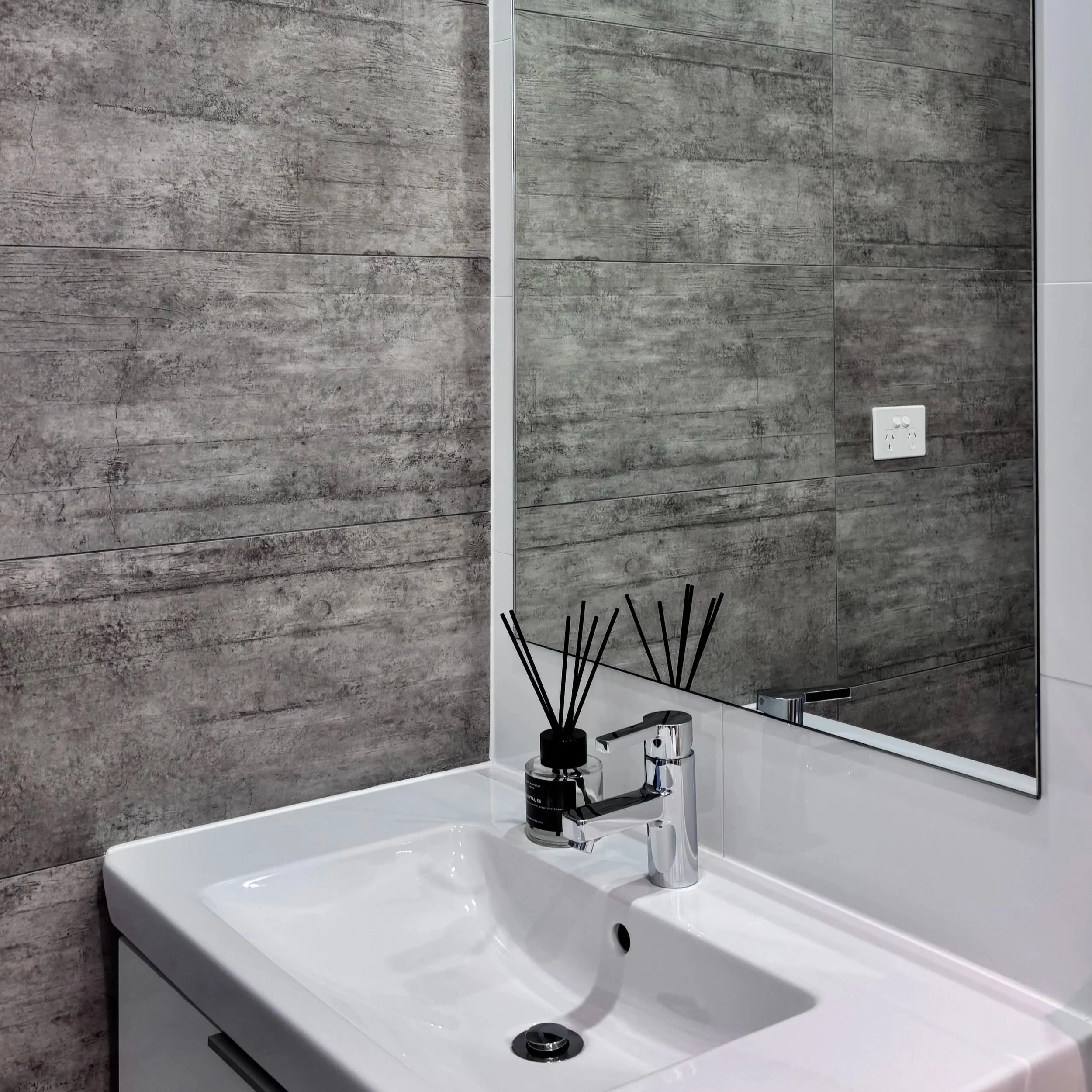 A bathroom sink with a modern chrome faucet, black soap dispenser, and a black container with reed diffuser sticks. The sink is installed on a white vanity, with a large mirror above it. The wall behind has gray wood-like textured tiles, and an elect