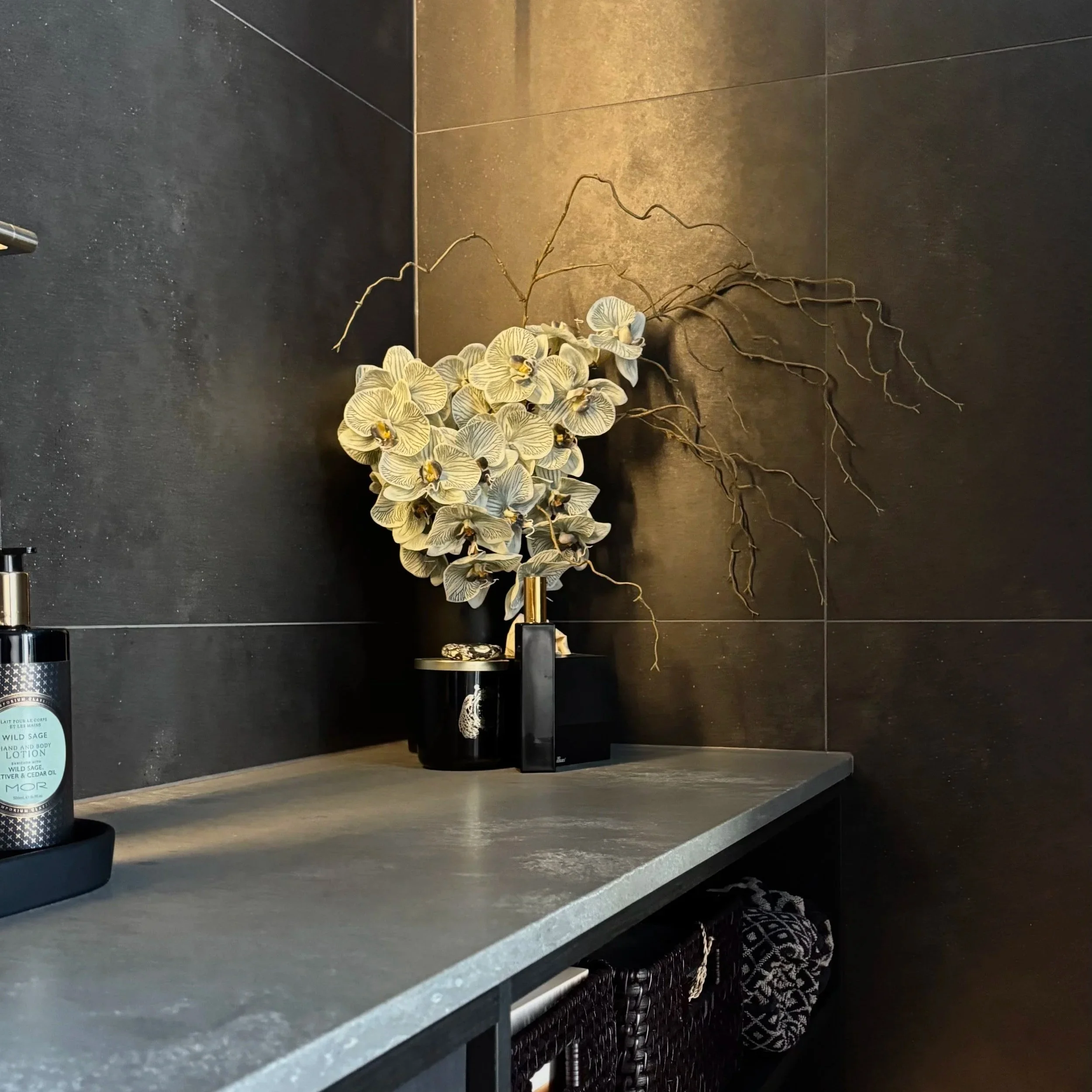 A dark kitchen countertop with a potted orchid and a lotion bottle, with black tiles on the wall above.