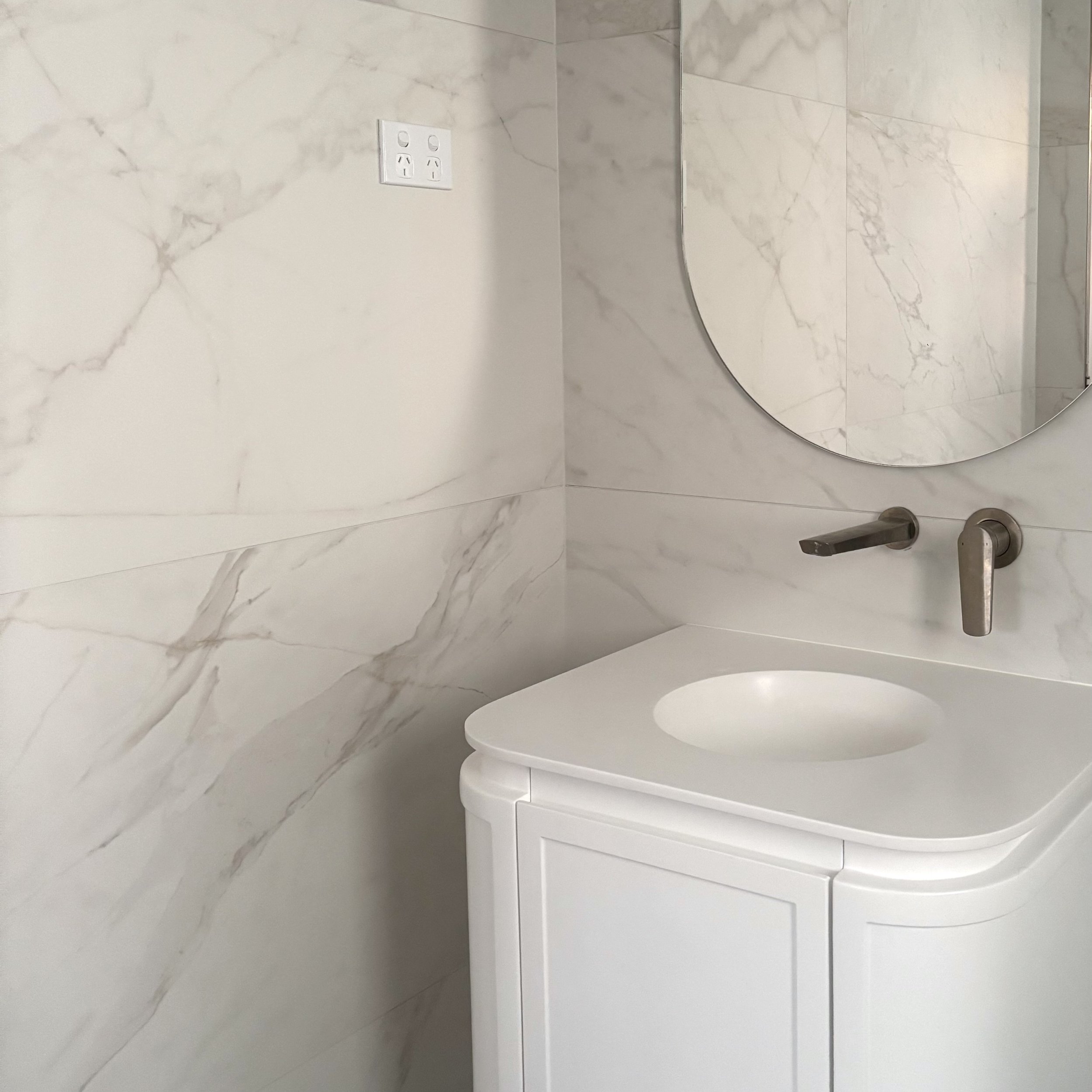 White marble bathroom wall with electrical outlets, a mirror, and a white toilet with a closed lid, seen from an angle.