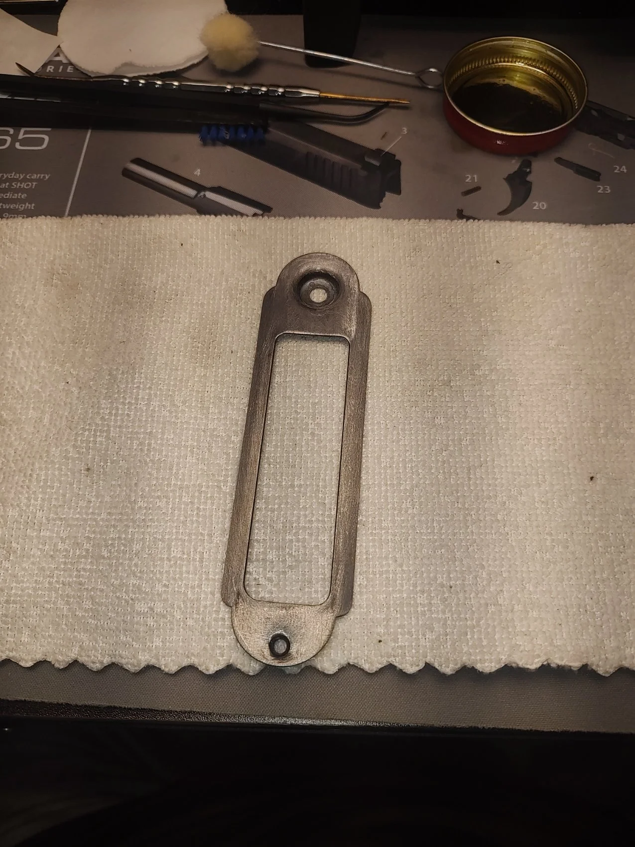 A metallic part, possibly from a firearm, placed on a white cloth. The background shows tools and a small container. The part has a rectangular opening and two holes at the ends.