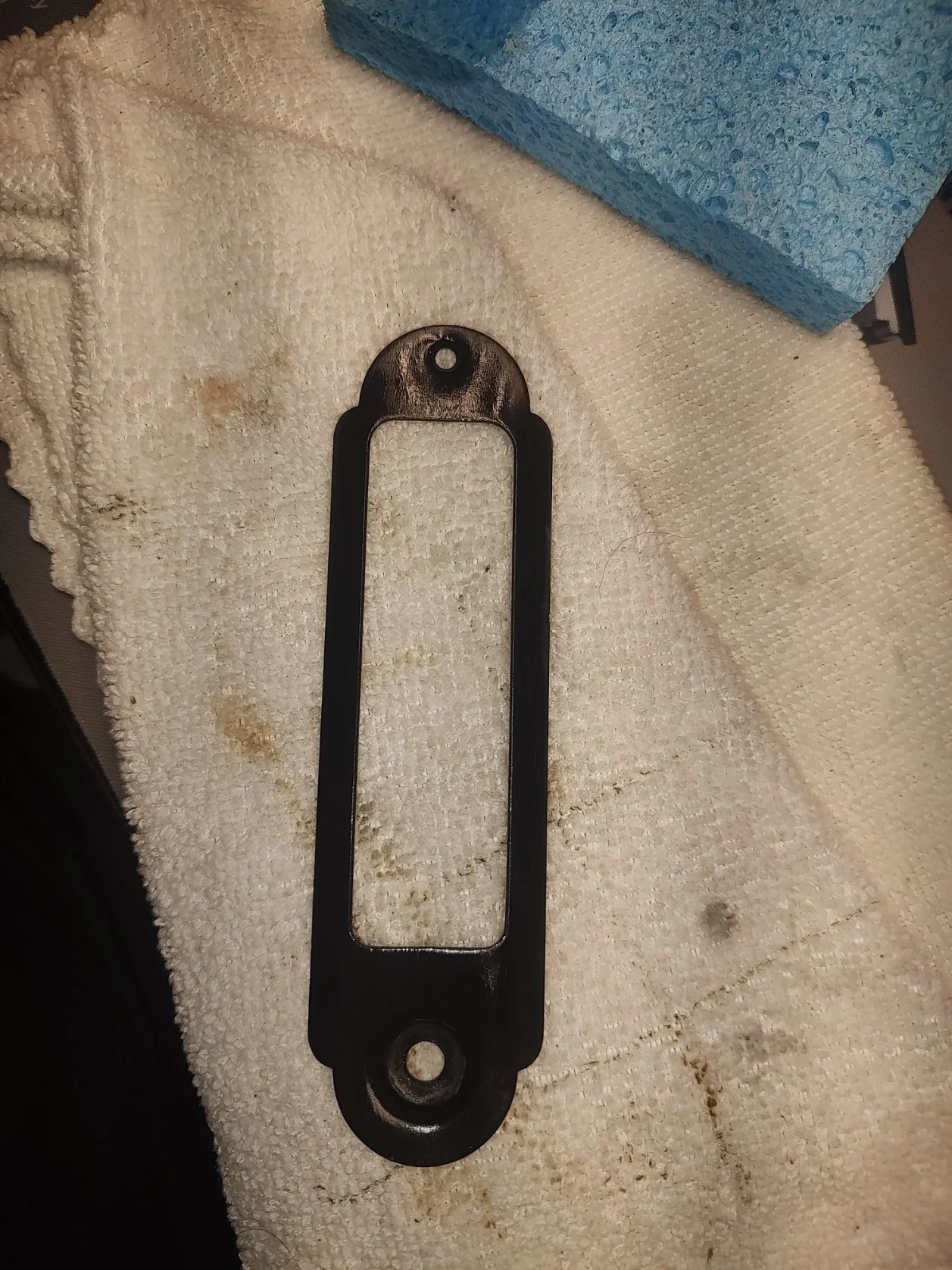 A black metal tool with a rectangular central opening and two circular ends, placed on a dirty white cloth with some brown stains.