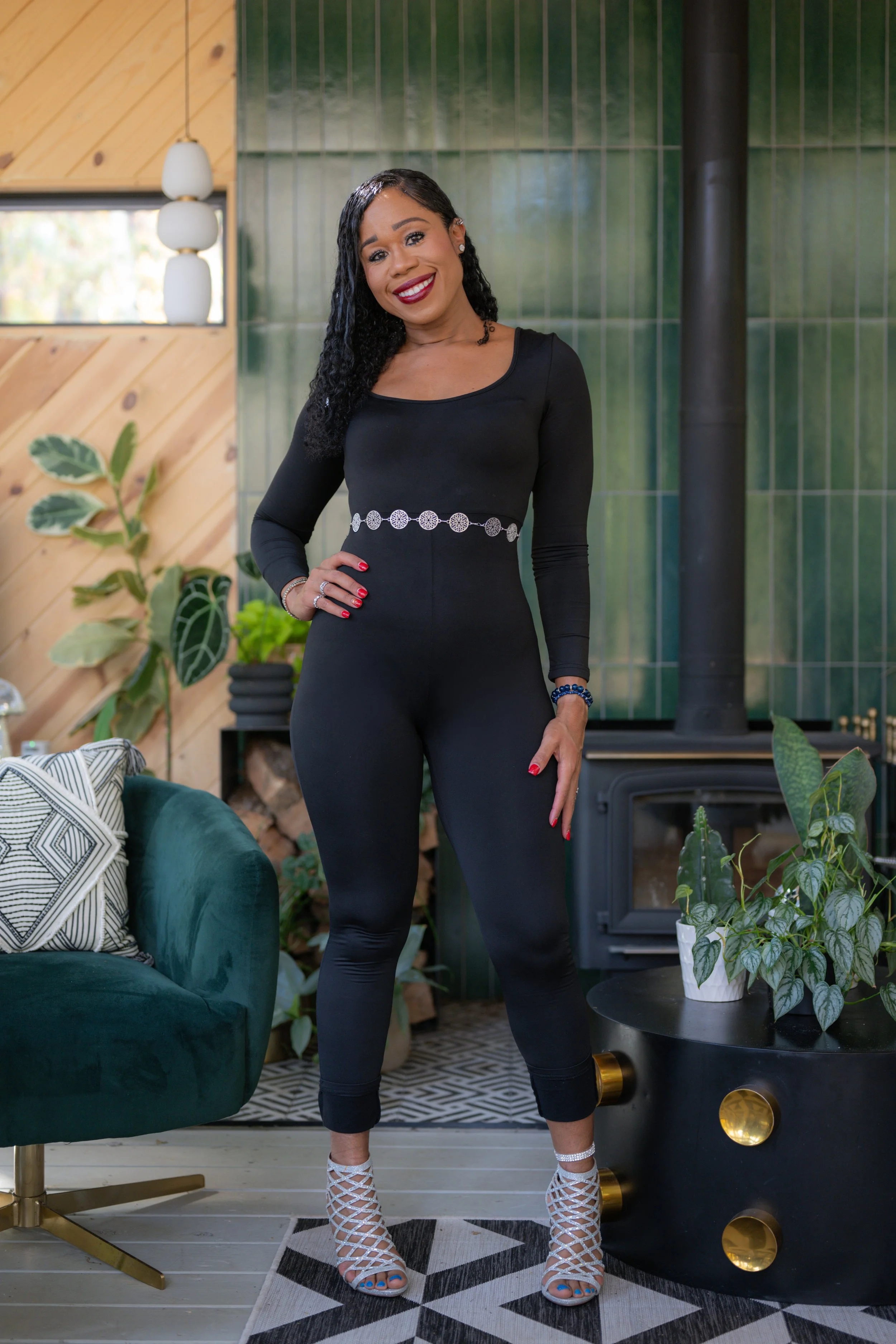 Woman in black jumpsuit standing in modern living room with plants, fireplace, and patterned rug.