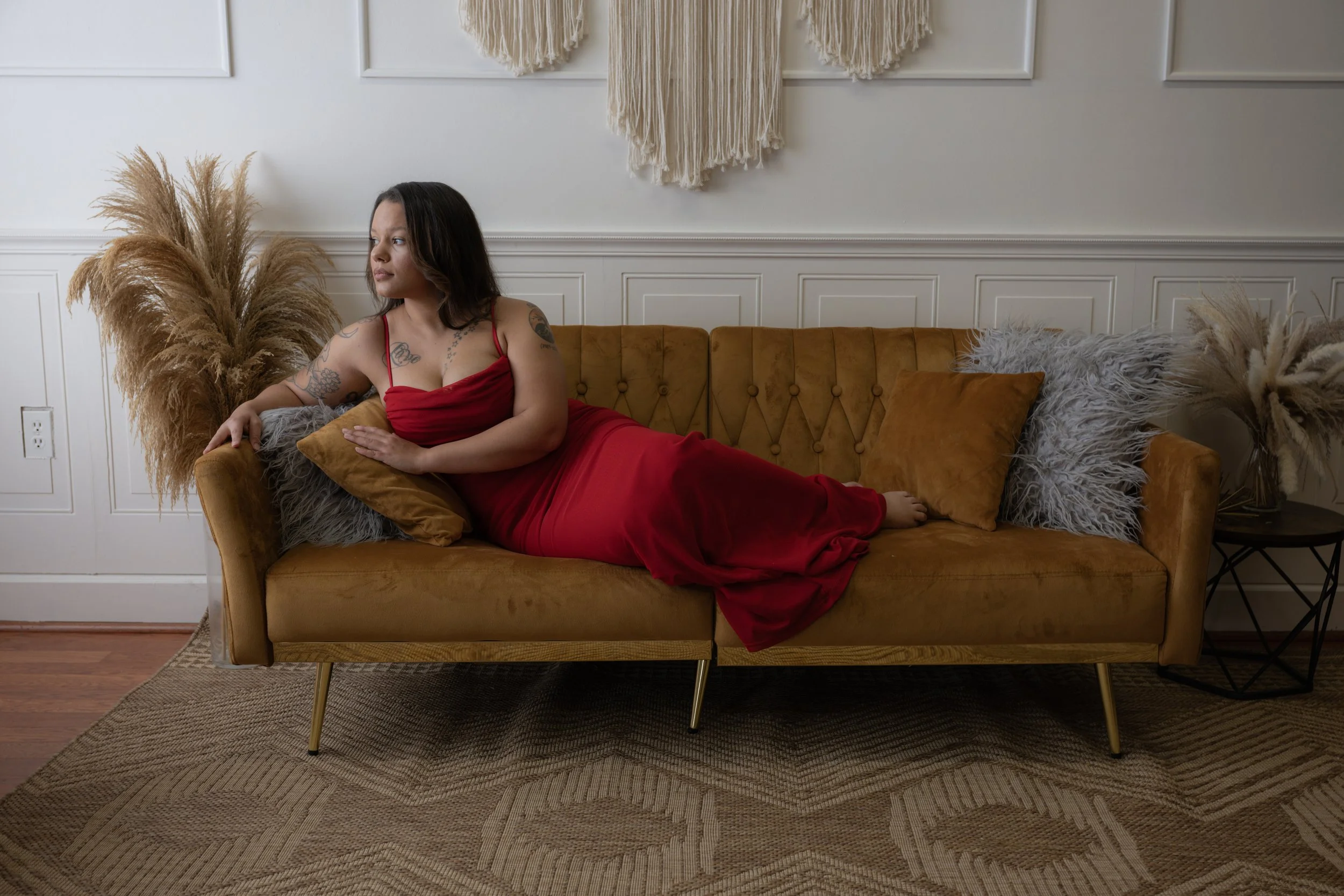 A woman lying on a mid-century modern brown velvet sofa in a living room, wearing a red dress, with decorative pillows and plants around, facing left with her arm resting on a pillow.
