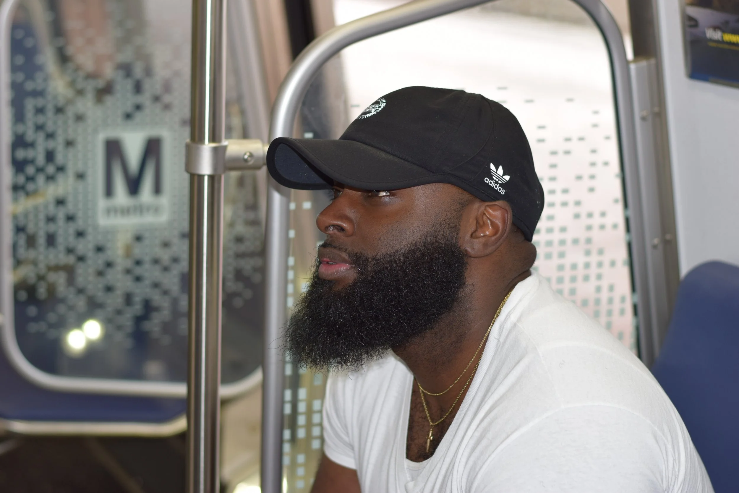 A man with a dark beard wearing a black Adidas cap and a white t-shirt, sitting on a bus or train with a metallic handlebar and a window in the background.