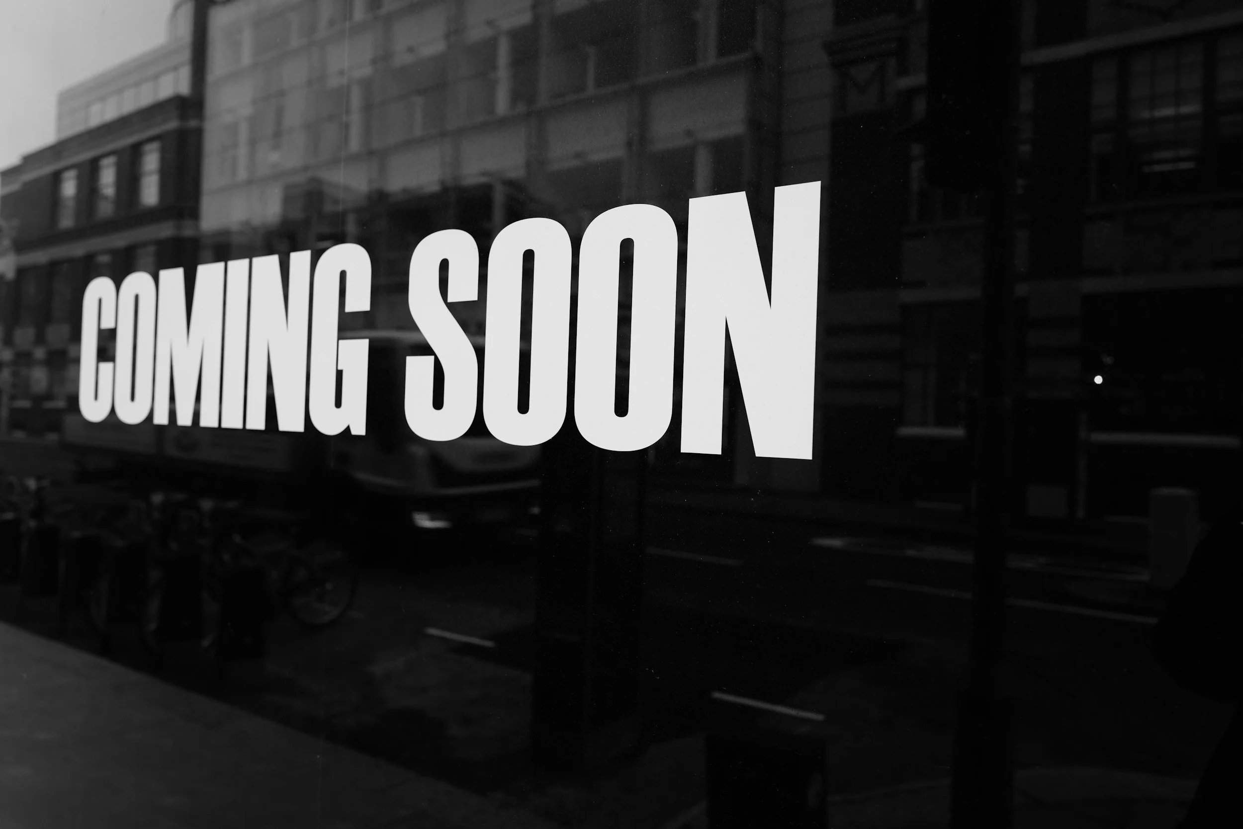 Black and white reflection of buildings and street in window with the words 'Coming Soon' in bold white letters.