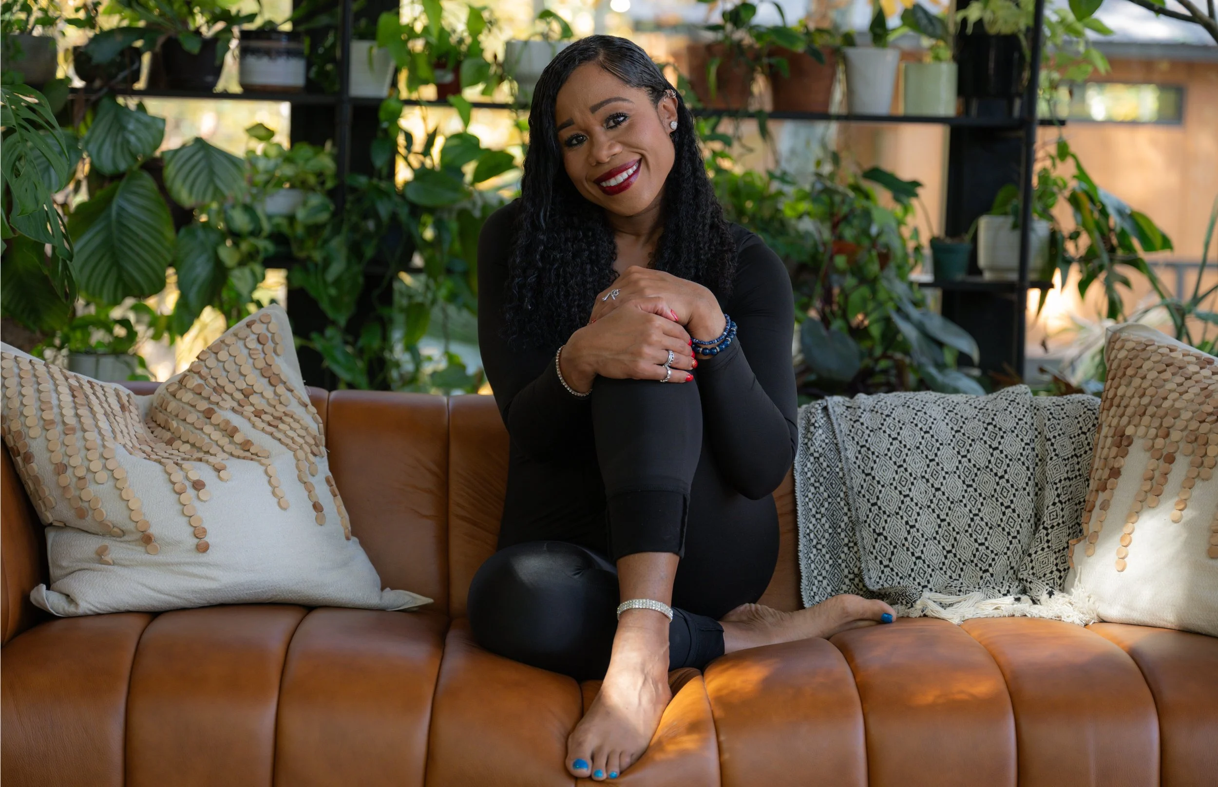 Smiling woman in black outfit sitting barefoot on a brown leather couch surrounded by decorative pillows, in a lush plant-filled indoor setting.