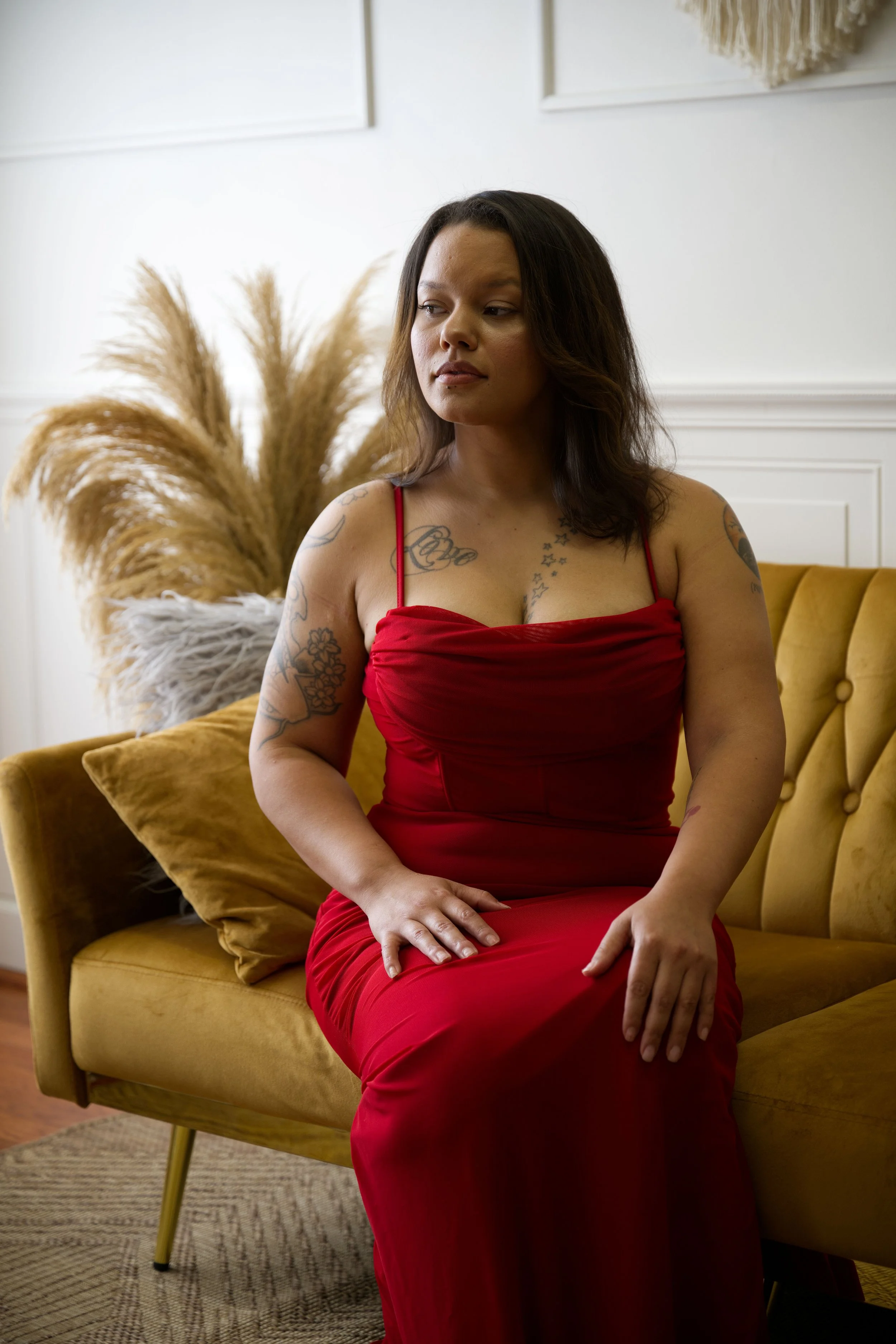 A woman in a red dress sitting on a mustard-colored couch in a room with white walls and decorative dried plants.
