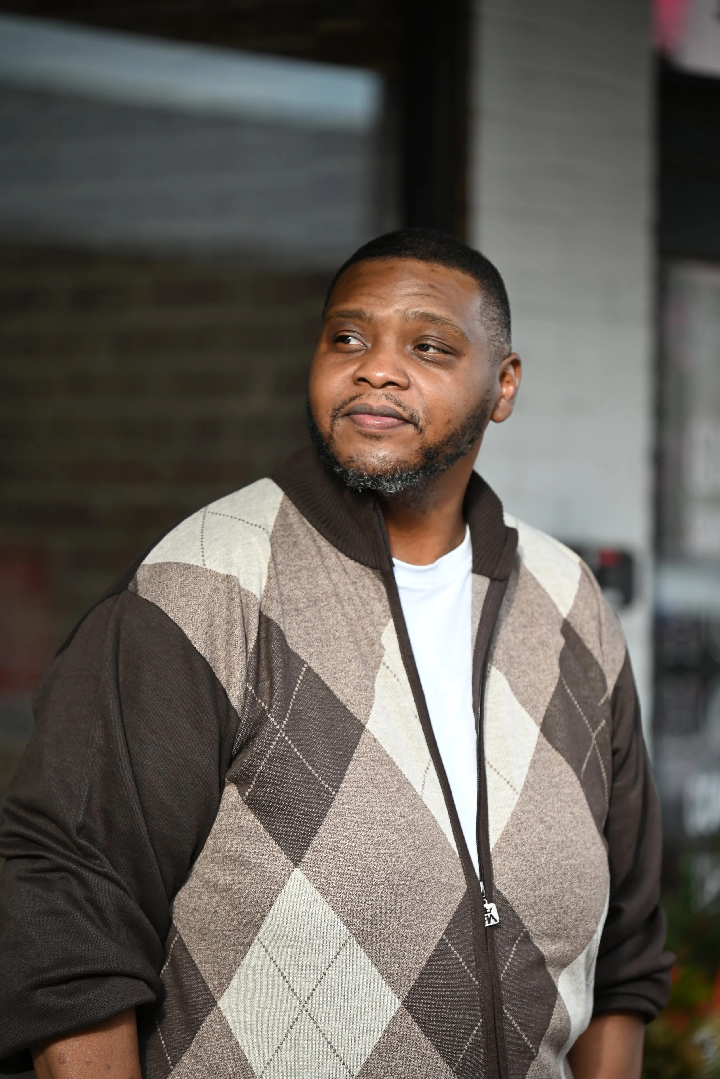 A man with short hair and a beard, wearing a argyle patterned jacket over a white t-shirt, standing outdoors with a relaxed expression.